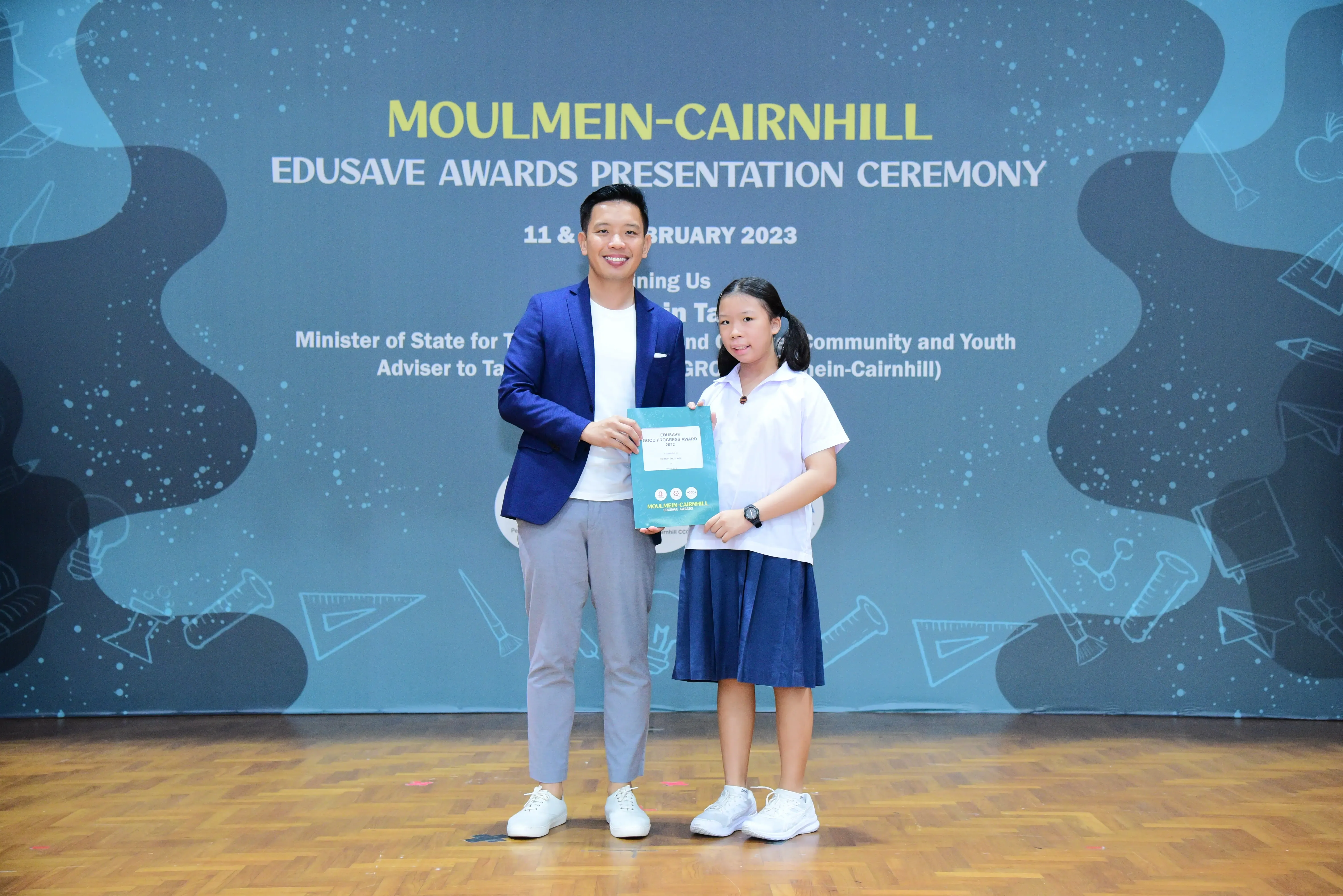 A man in a blue blazer and a young girl in school uniform hold a certificate at the Moulmein-Cairnhill Edusave Awards 2023.