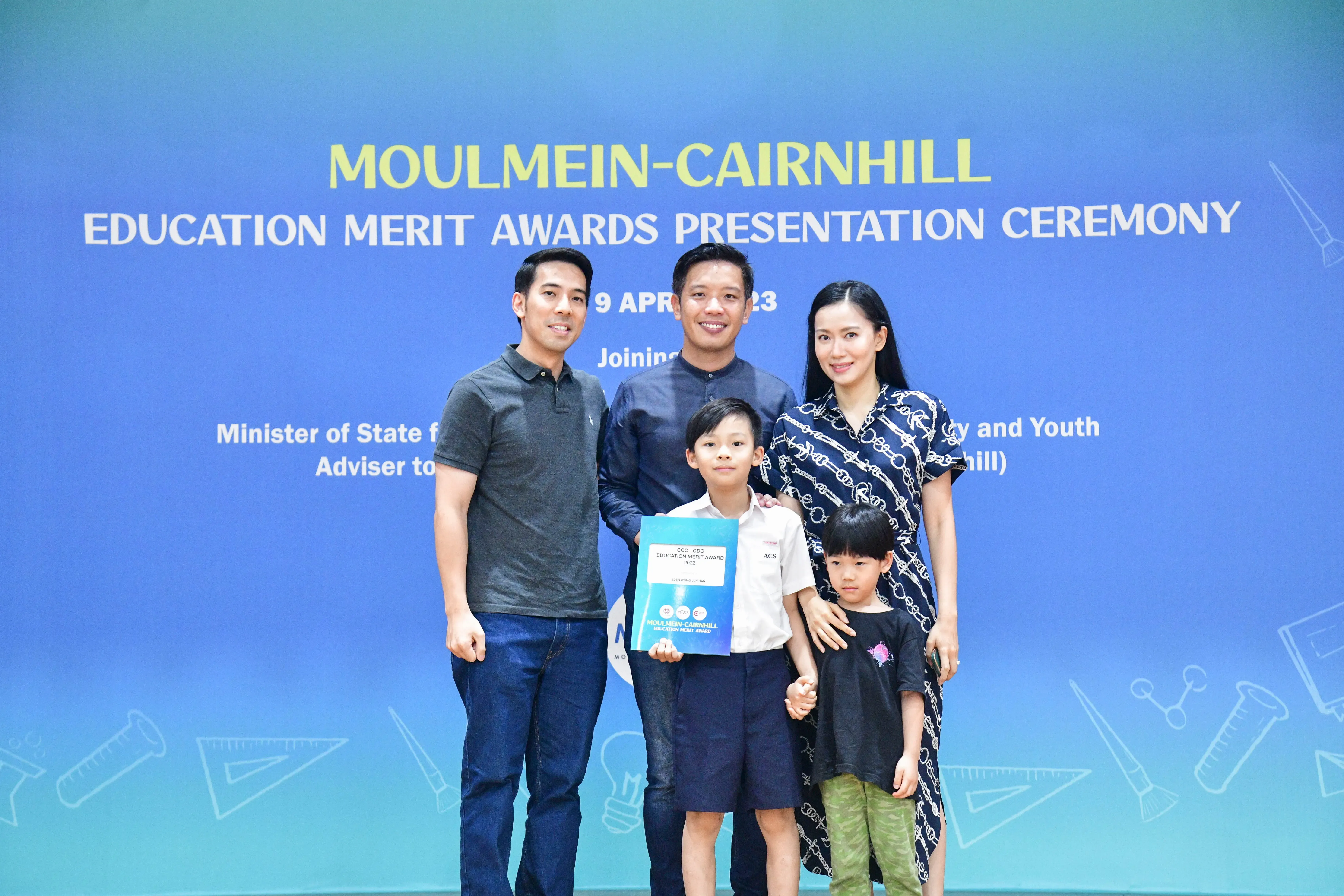 Three adults and two children pose before a Moulmein-Cairnhill Education Merit Awards Presentation Ceremony banner.