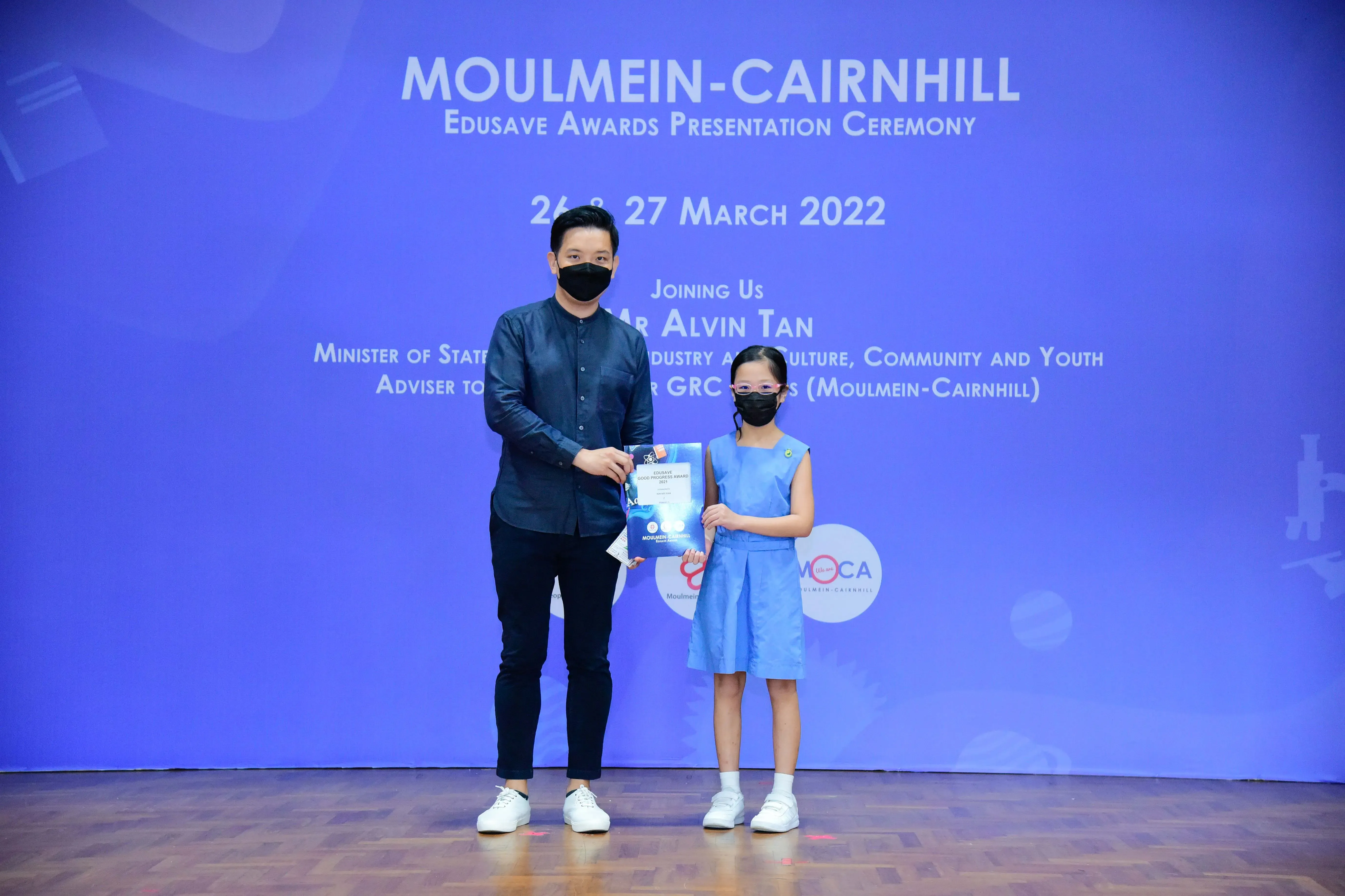 An adult and a young girl in masks hold a certificate at the Moulmein-Cairnhill Edusave Awards Ceremony, March 2022.