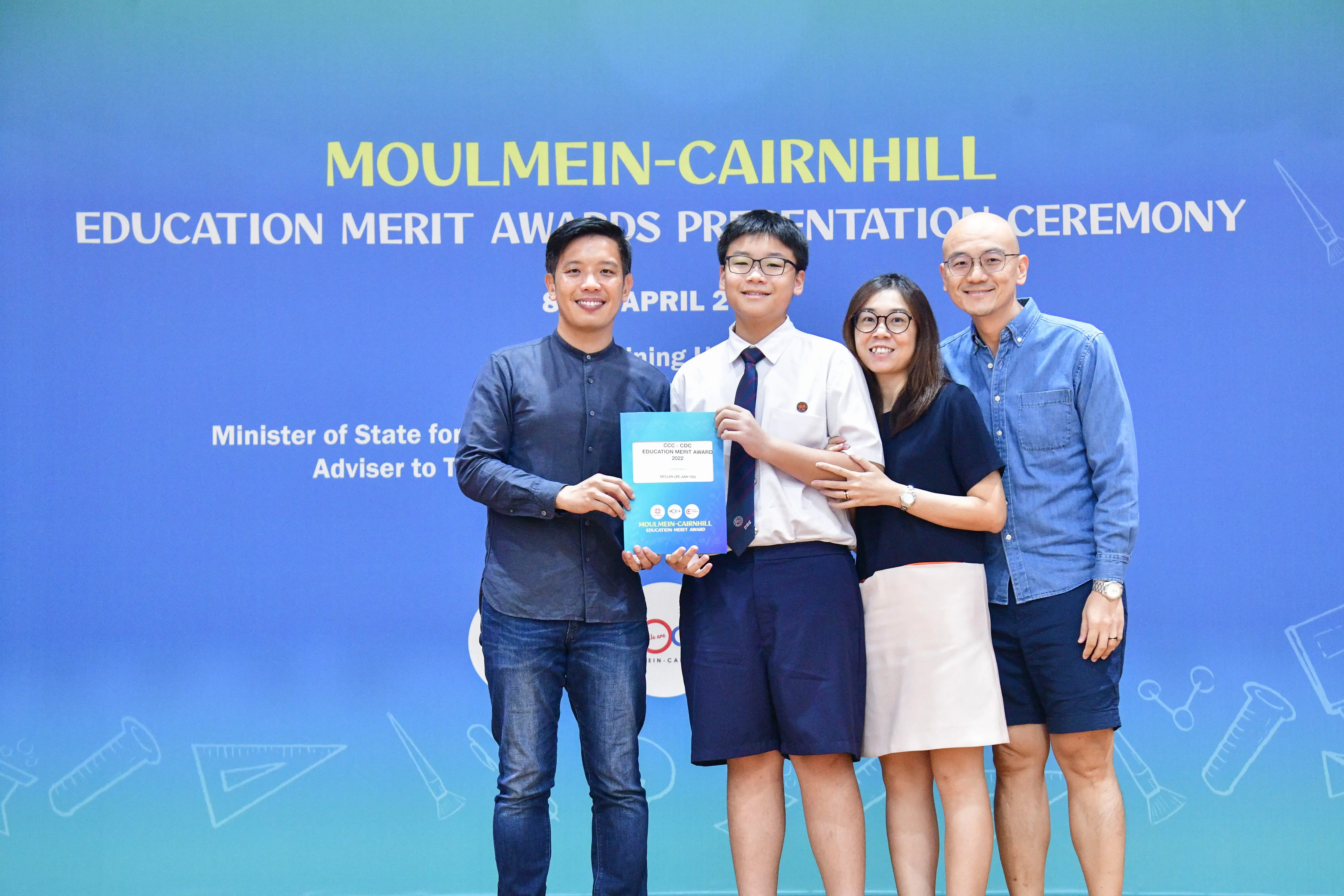 Four people smile at the Moulmein-Cairnhill Education Merit Awards Presentation Ceremony, with a student holding an award.