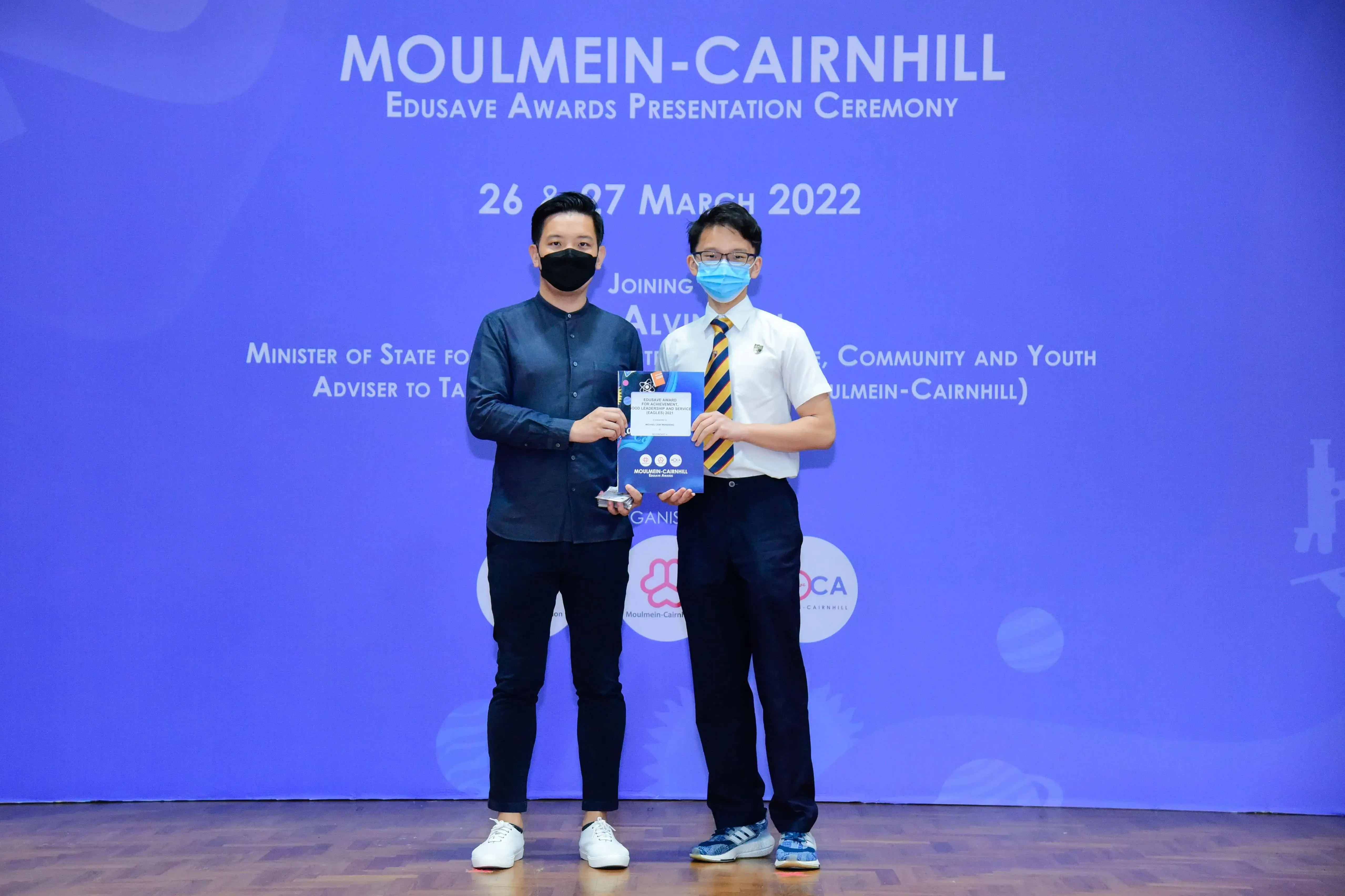 Two masked men hold an award at the Moulmein-Cairnhill Edusave Awards Presentation Ceremony, March 2022.