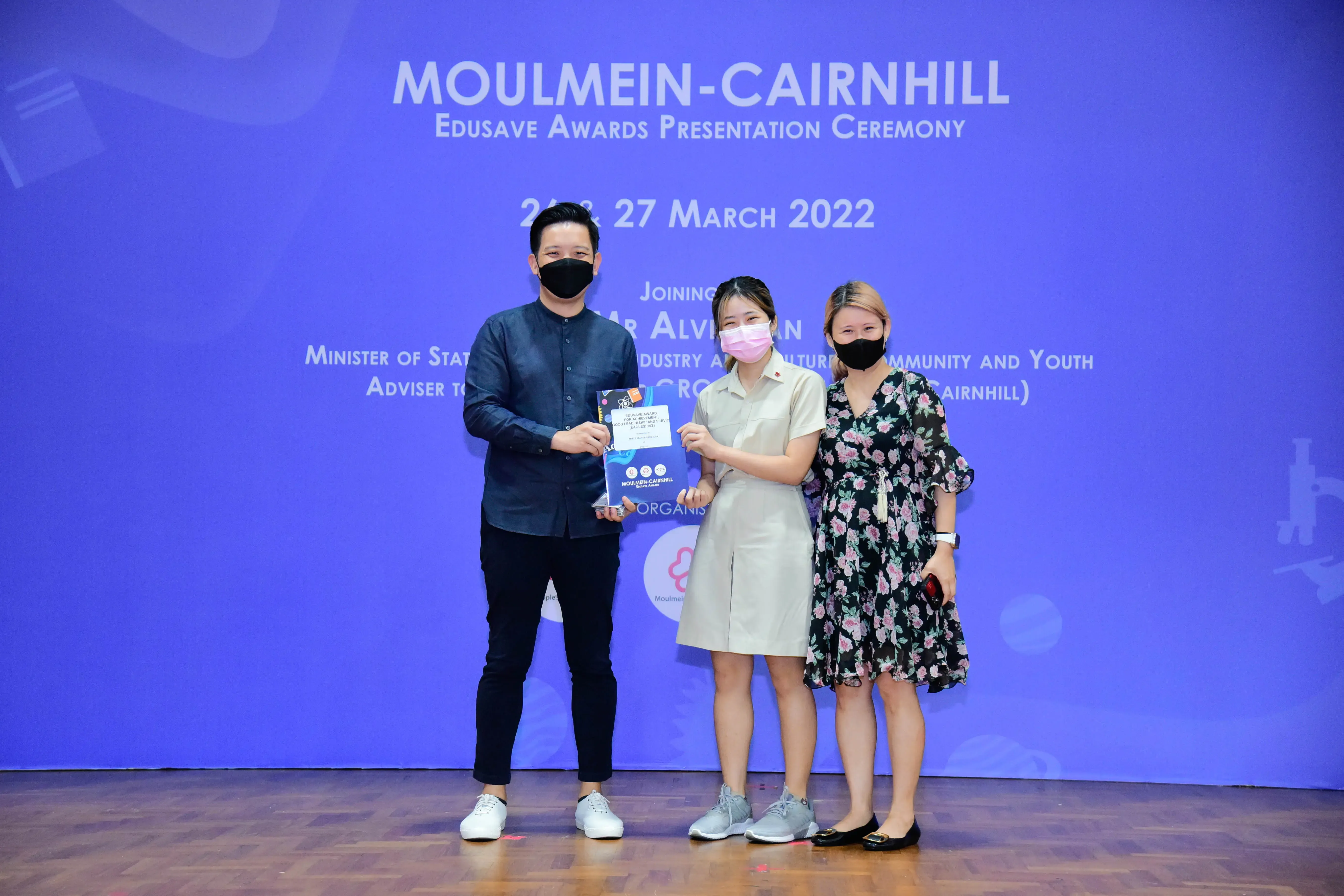 Three masked people pose holding an award booklet at the Moulmein-Cairnhill Edusave Awards Ceremony, March 2022.