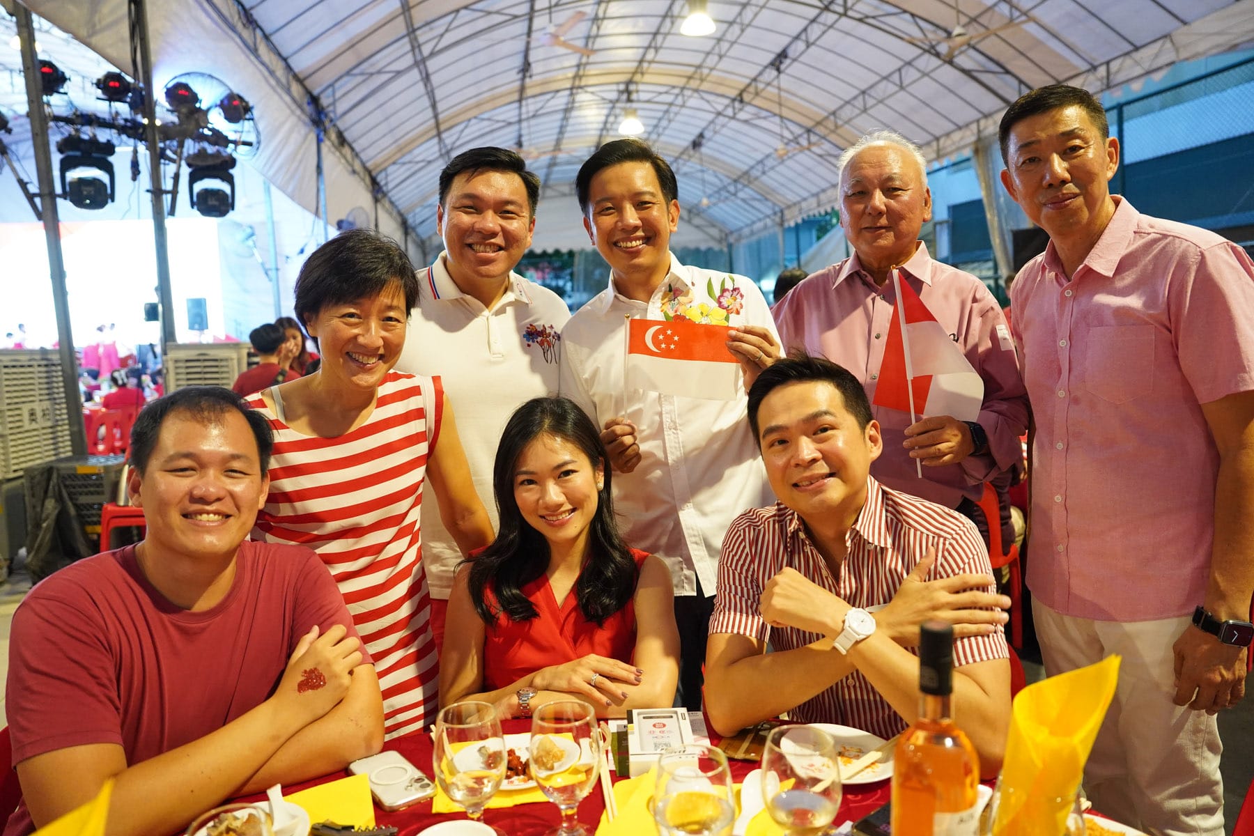 A group of eight smiling people in red and white outfits pose at a festive table, holding small Singapore flags under a tent.