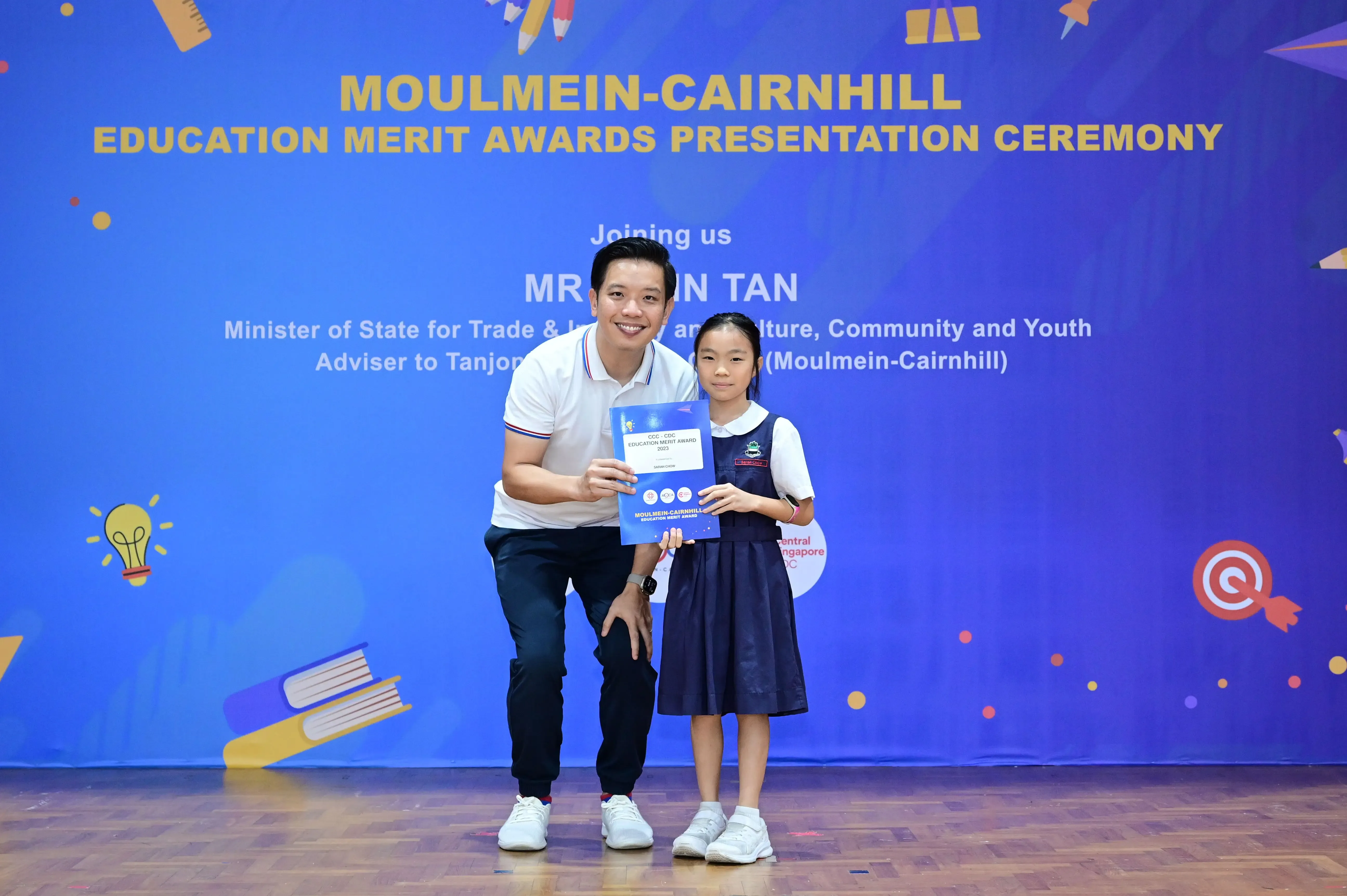 A man and a young girl in school uniform hold an award certificate in front of a Moulmein-Cairnhill Education Merit Awards banner.