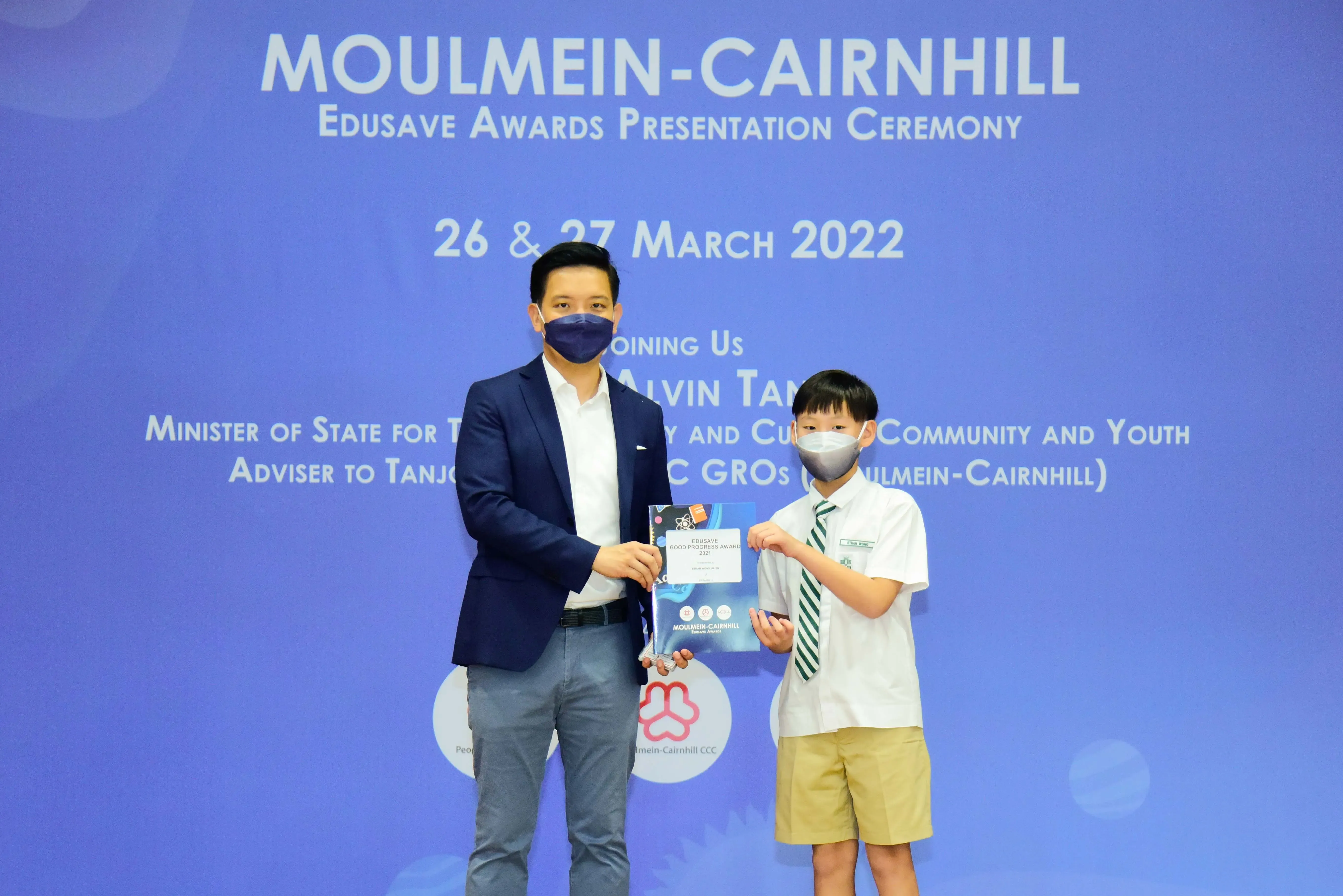 An adult in a blue blazer presents an award certificate to a young student at the Moulmein-Cairnhill Edusave Awards Ceremony.