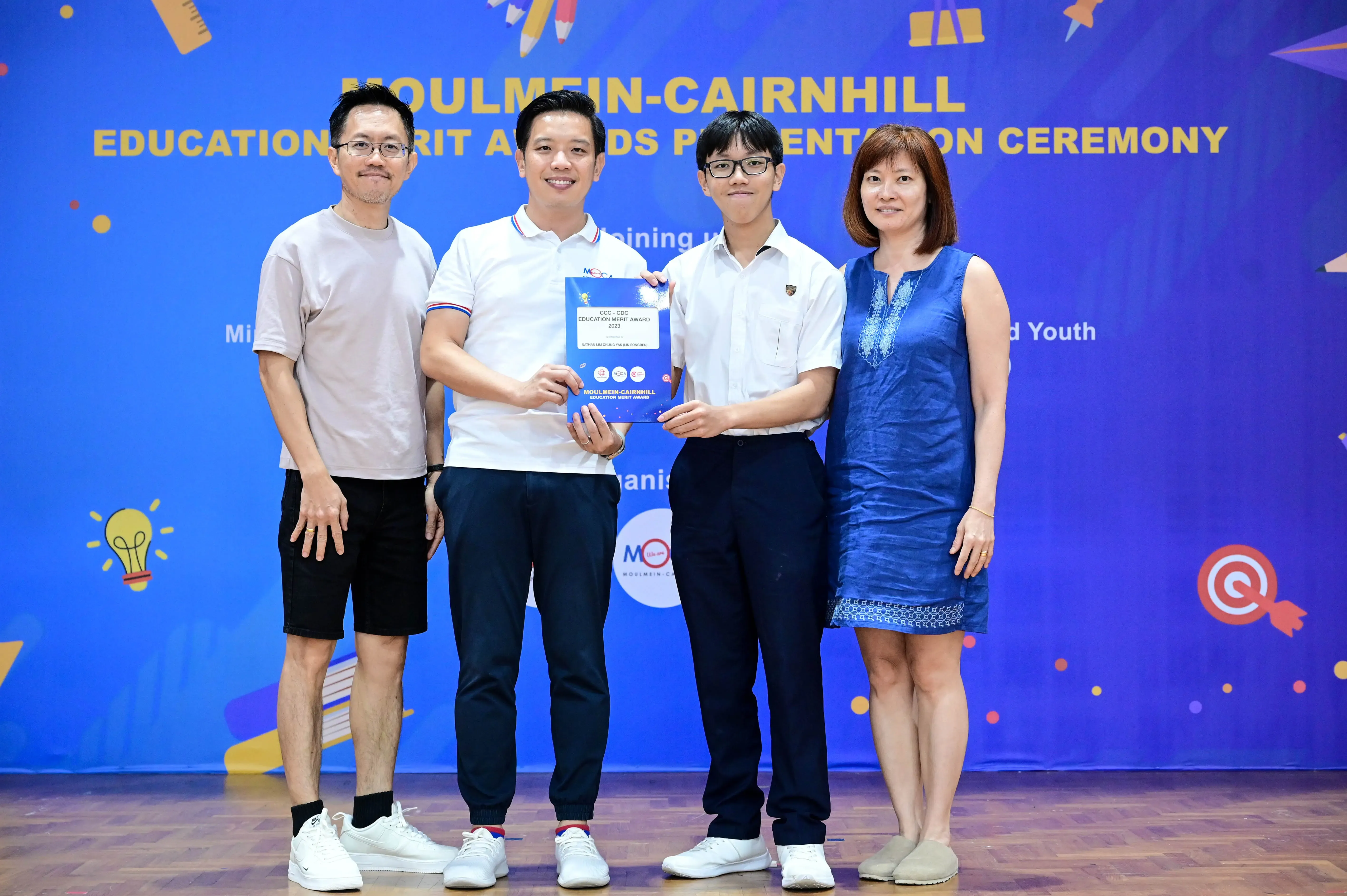 Four people stand before a Moulmein-Cairnhill Education Merit Awards banner, two holding a blue award certificate.