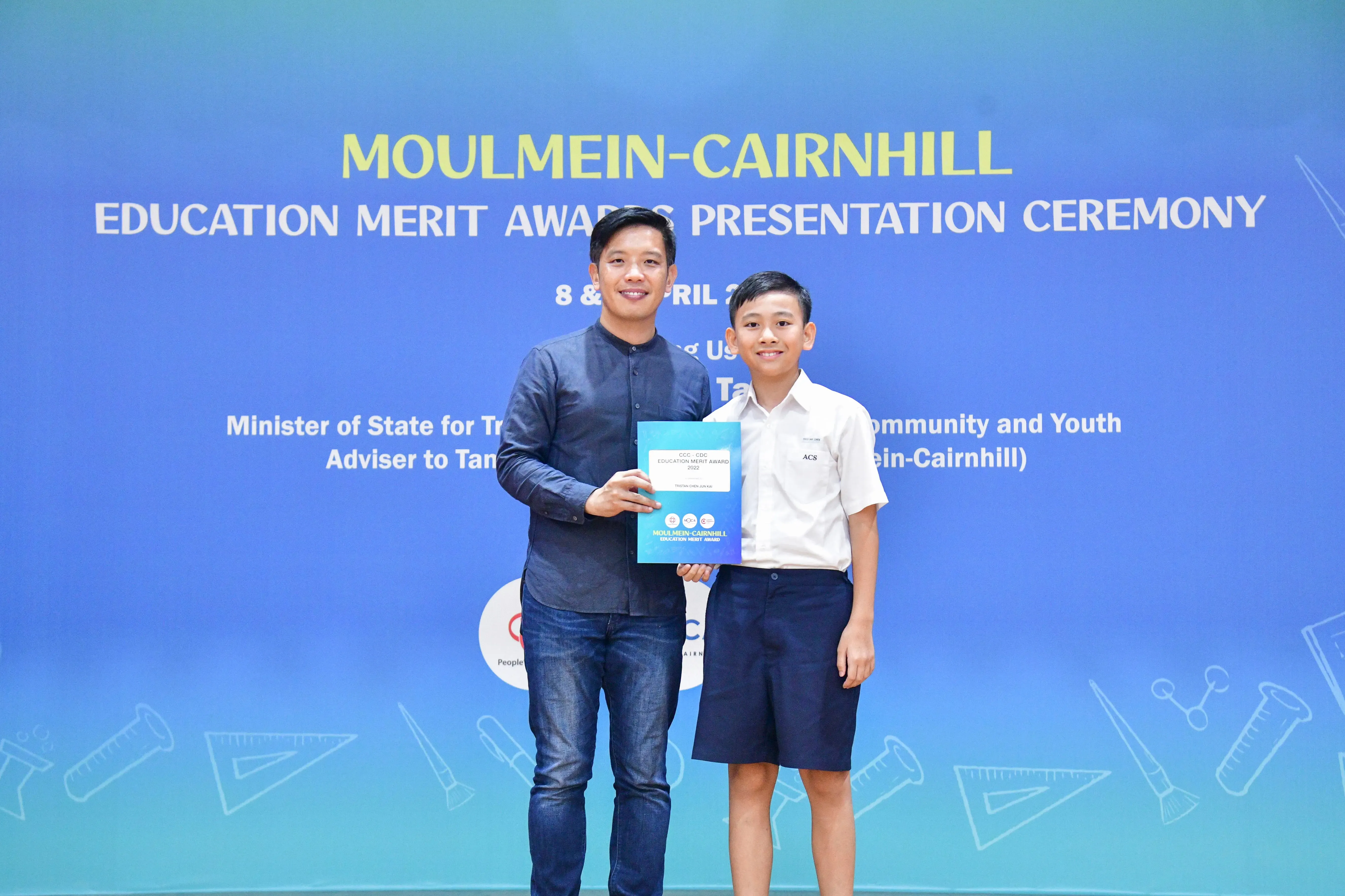 Adult and young boy holding a certificate together at Moulmein-Cairnhill Education Merit Awards ceremony.
