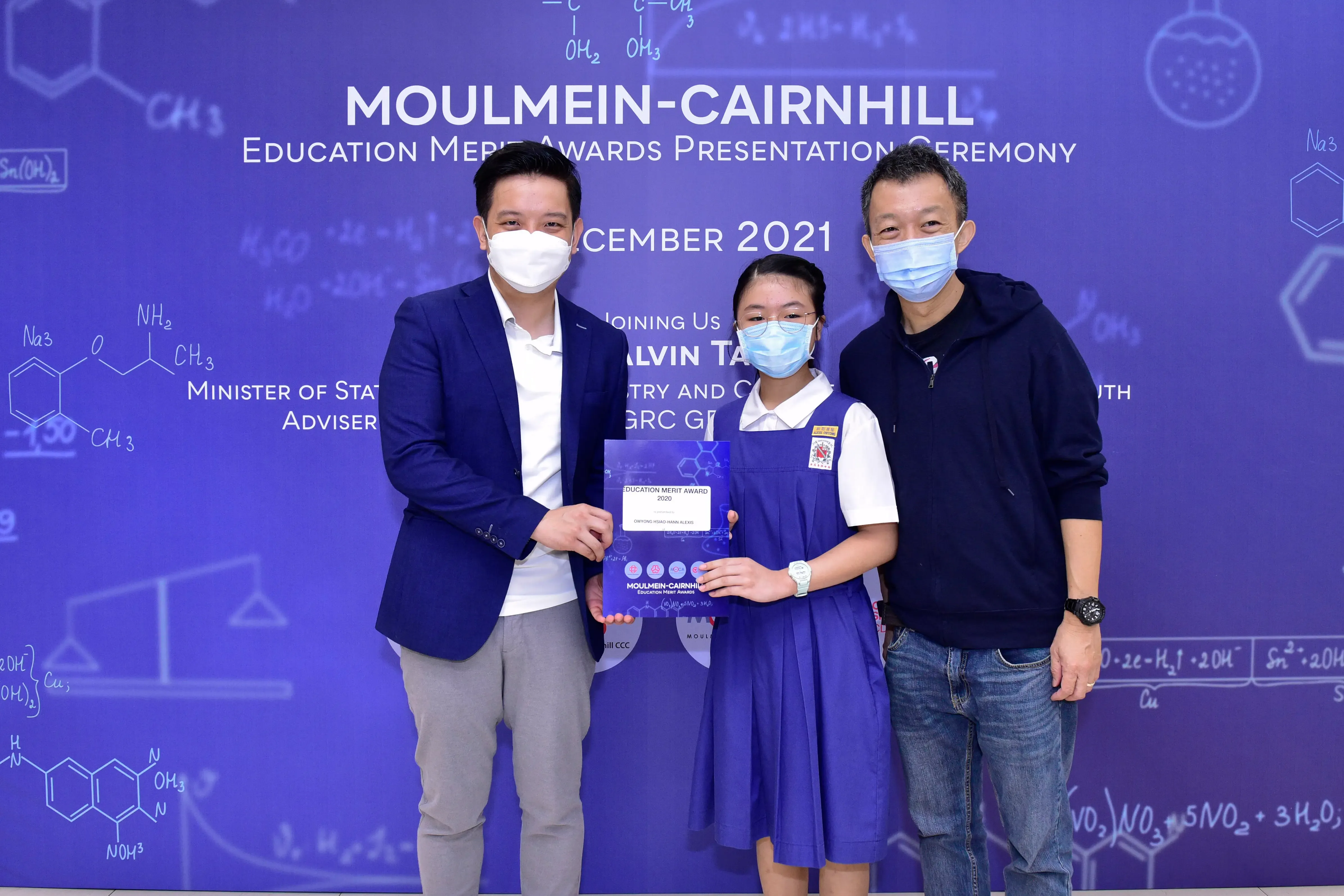 Three masked people pose at the Moulmein-Cairnhill Education Merit Awards Ceremony 2021, with a student holding a certificate.