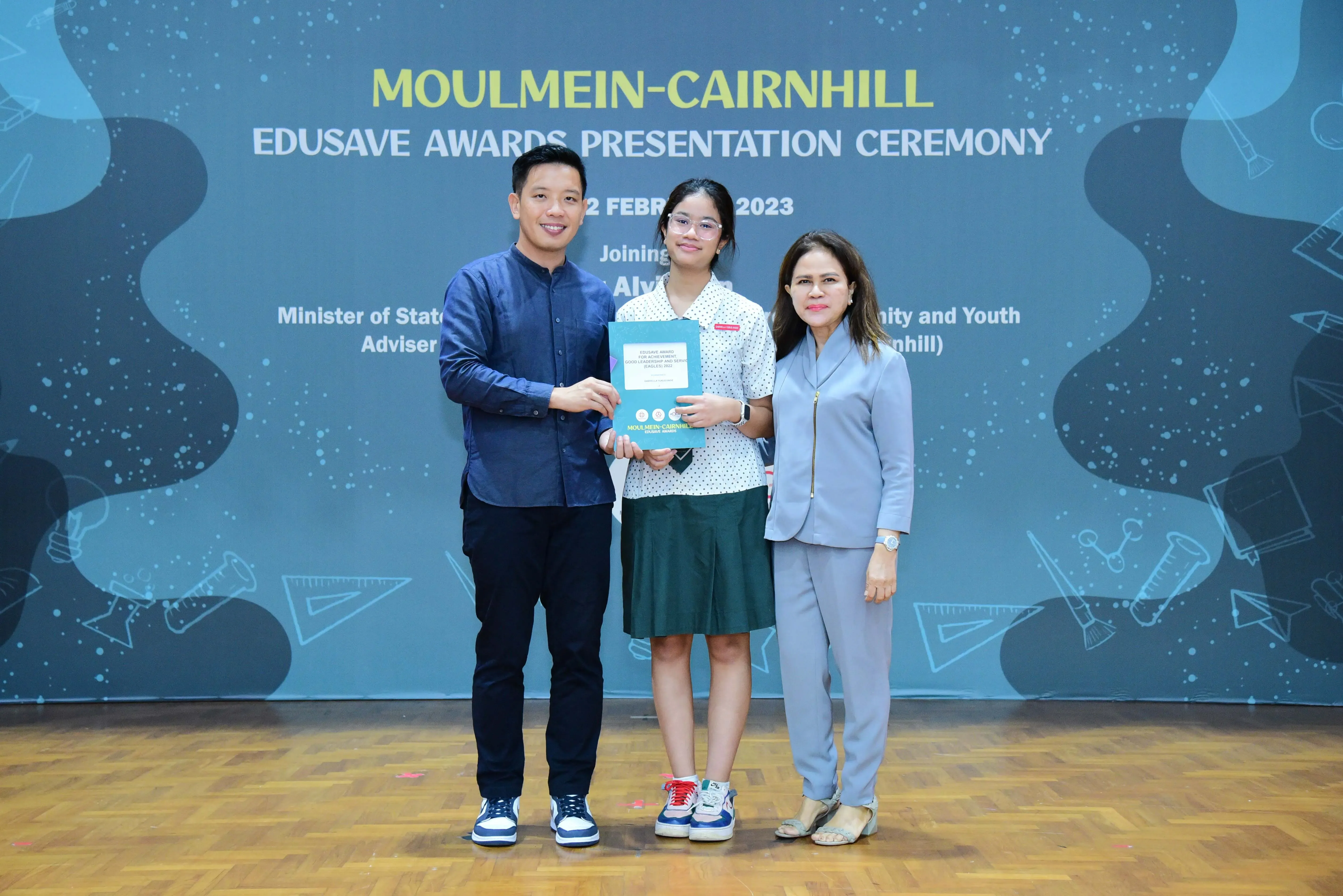 Three people pose on stage at the Moulmein-Cairnhill Edusave Awards Presentation Ceremony 2023, with a student holding an award.