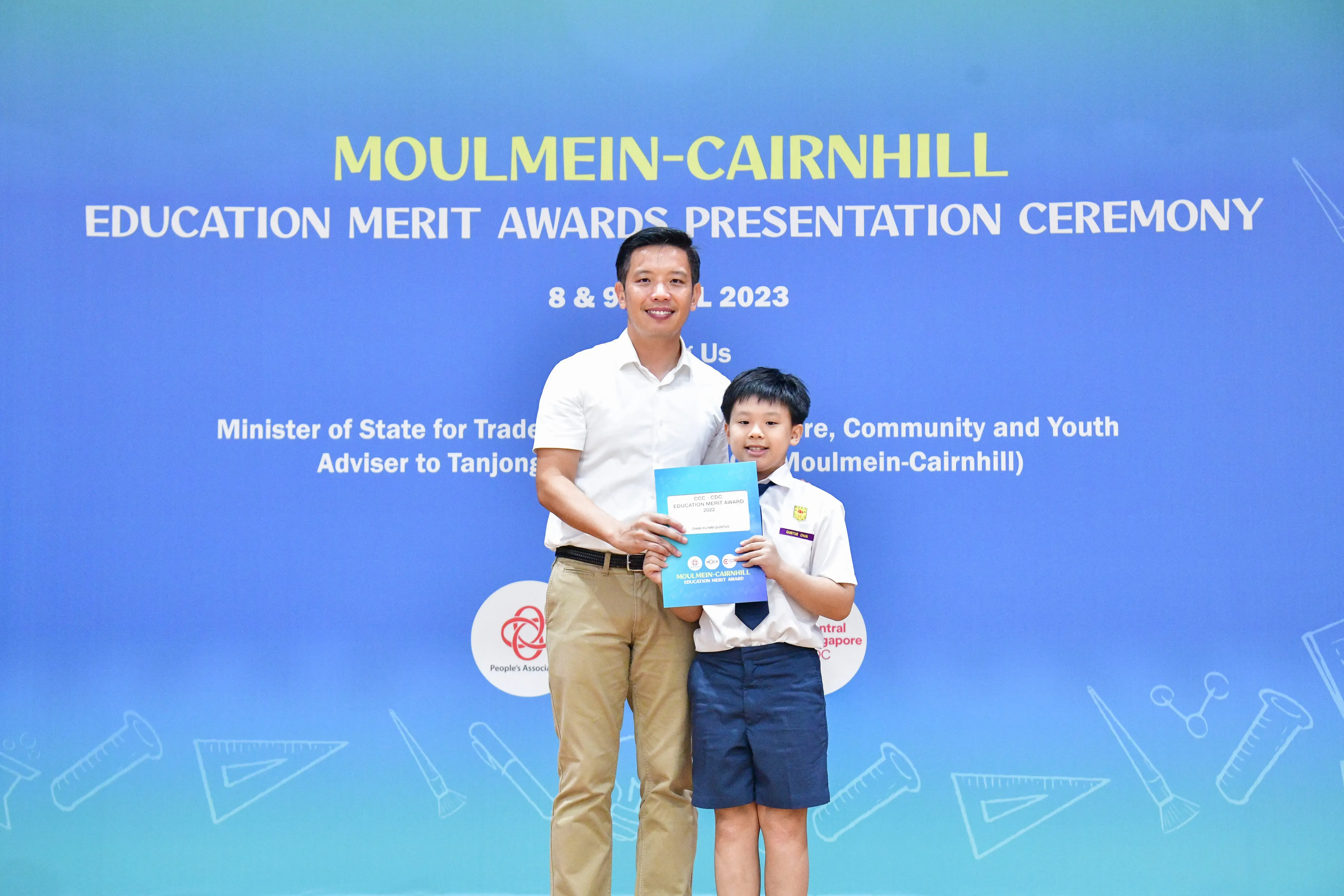 An adult man and a young boy in school uniform hold an award certificate at the Moulmein-Cairnhill Education Merit Awards 2023.
