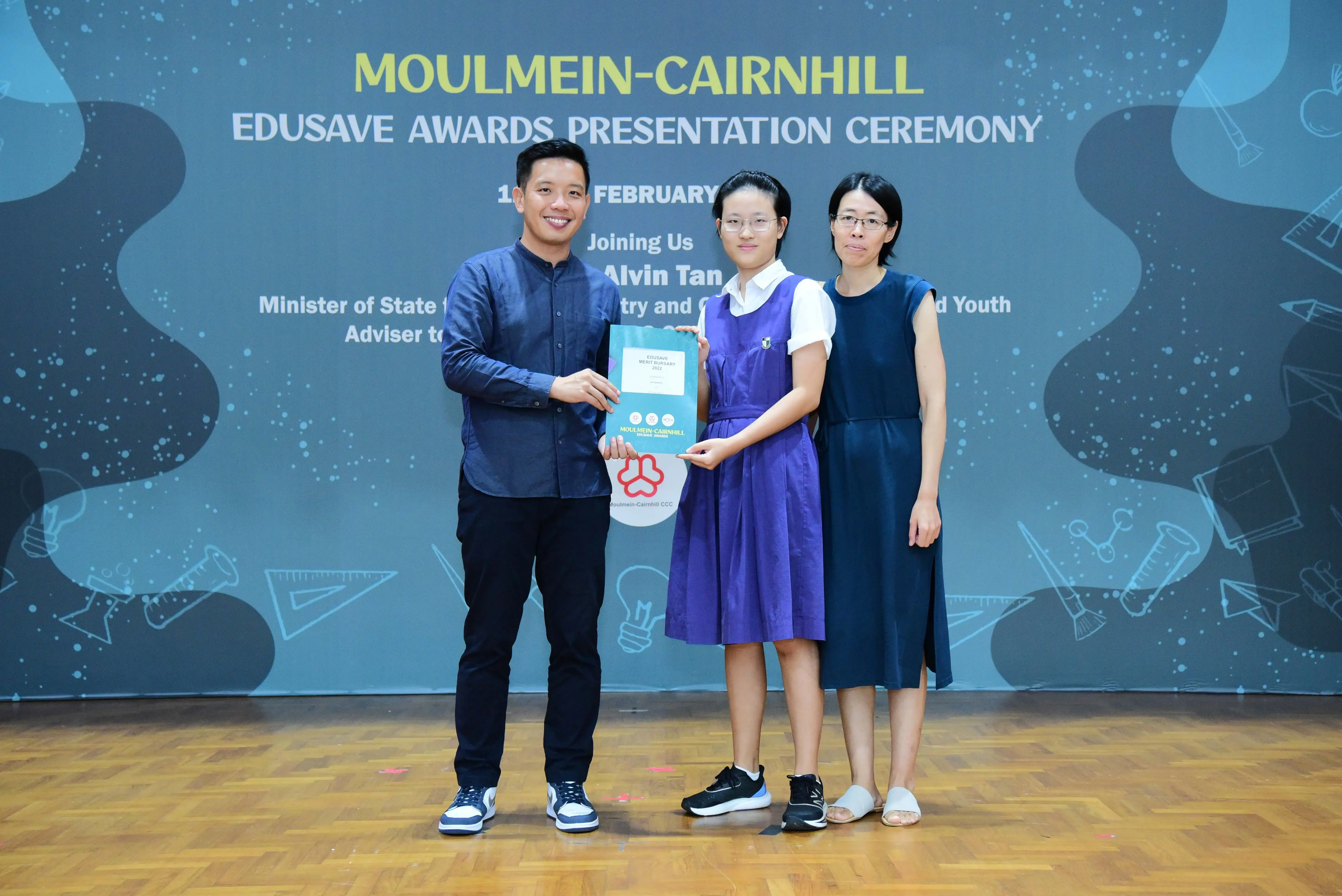 Three people pose holding an award booklet at the Moulmein-Cairnhill Edusave Awards Presentation Ceremony.
