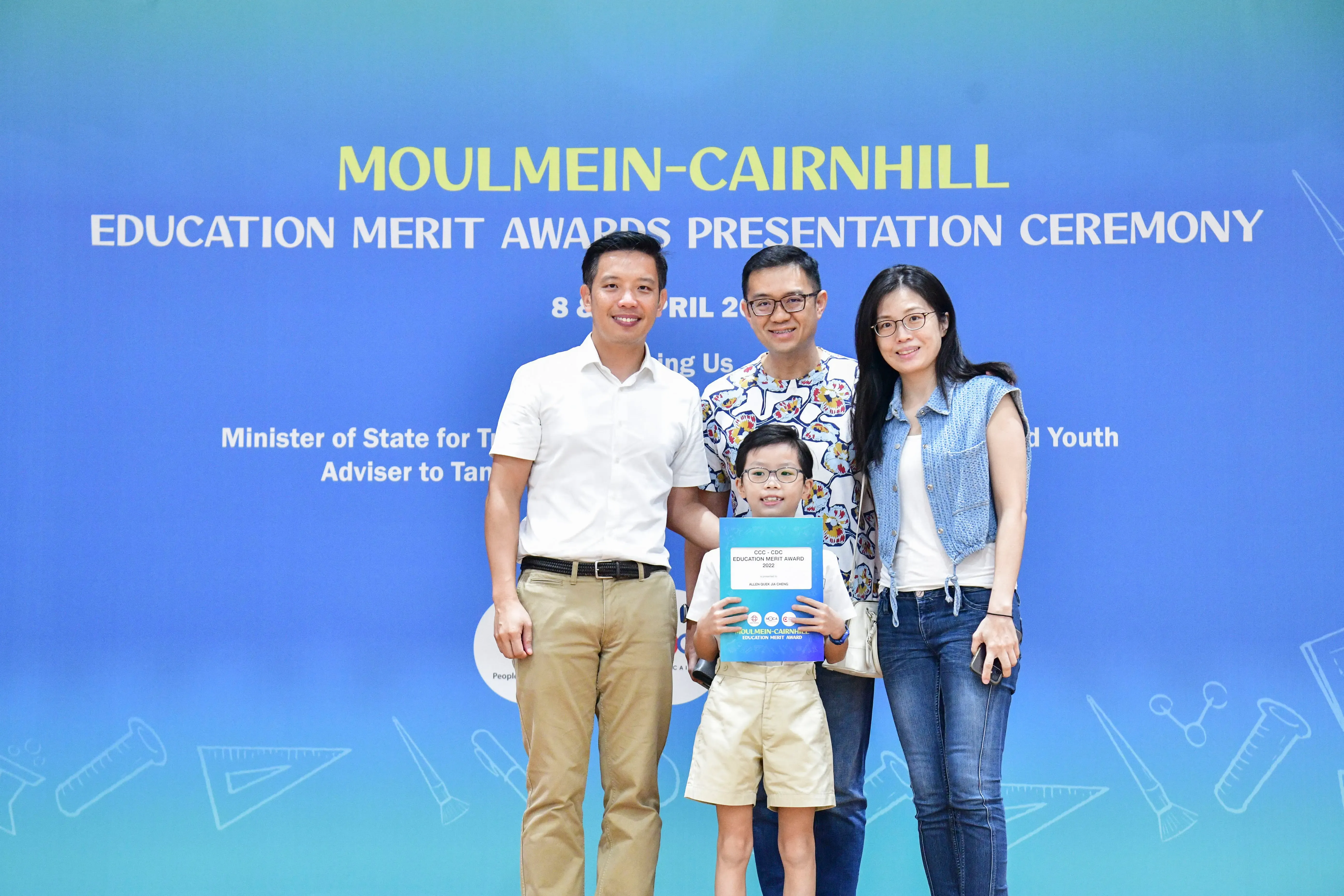 Three adults and a boy holding an award certificate pose in front of a Moulmein-Cairnhill Education Merit Awards banner.