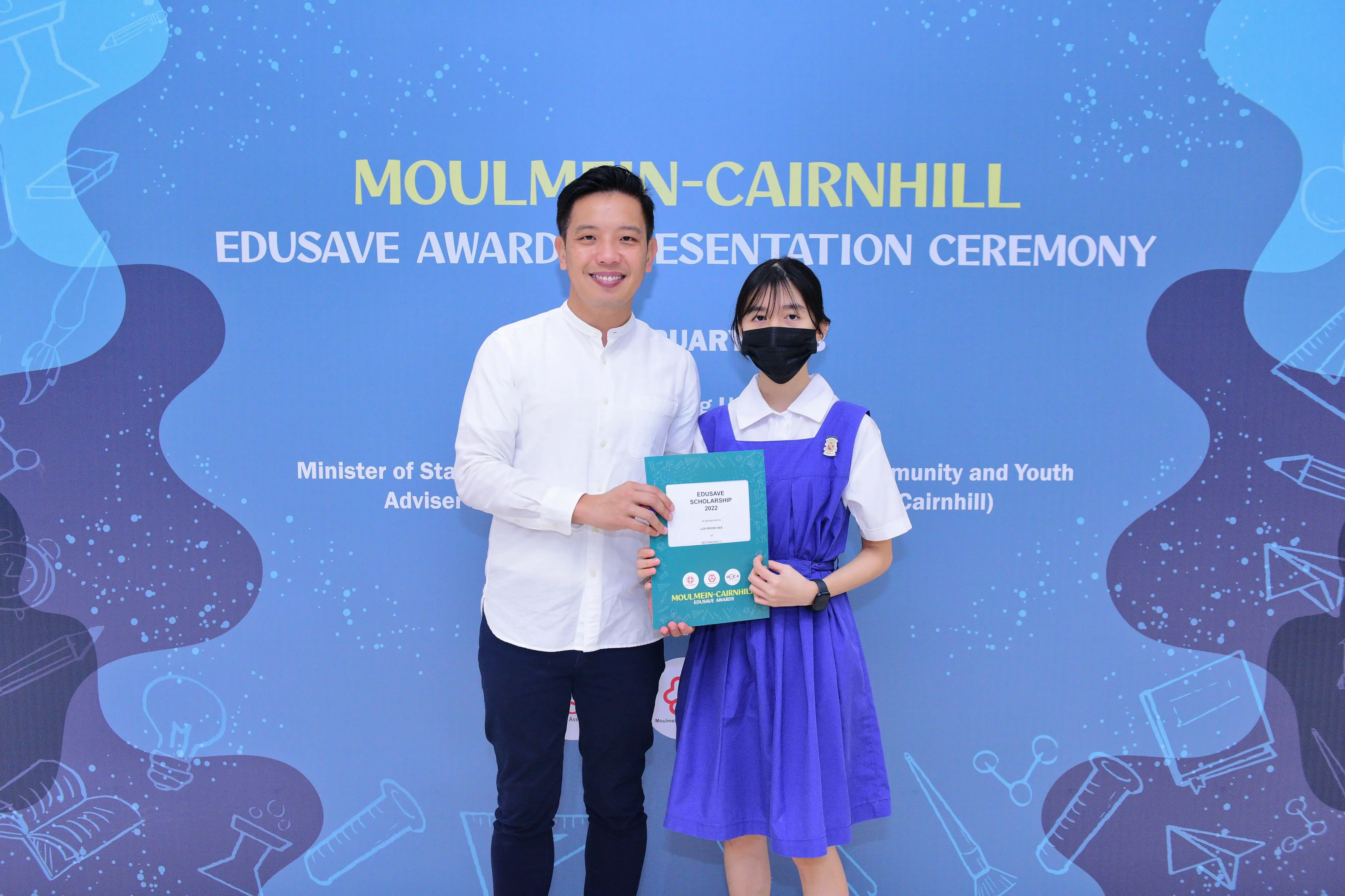 A man and a masked student hold an Edusave Scholarship certificate at the Moulmein-Cairnhill Awards Ceremony.
