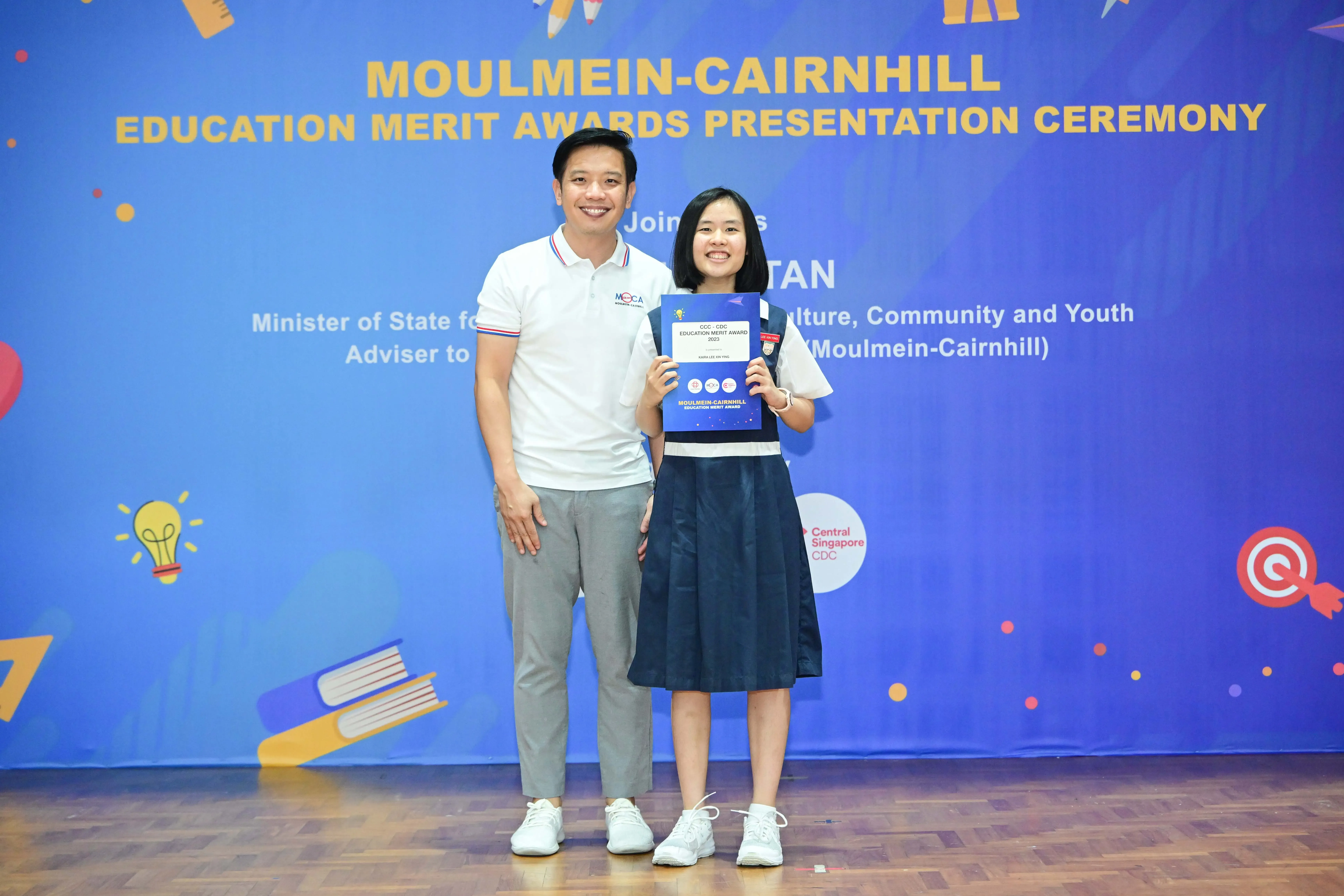 Two people smile on stage at the Moulmein-Cairnhill Education Merit Awards, girl holding a blue certificate folder.