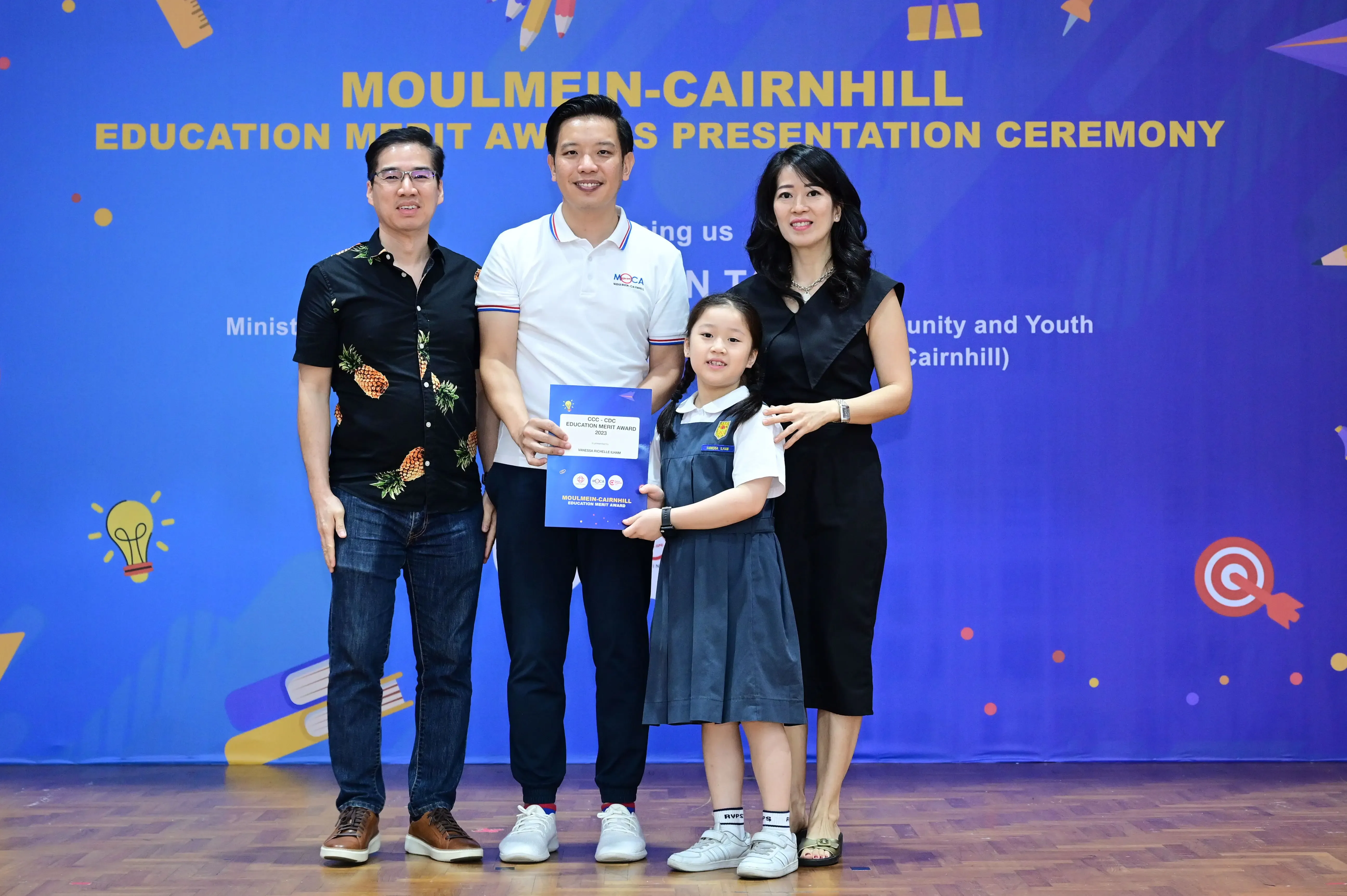 Three adults and a young girl in school uniform hold a certificate at the Moulmein-Cairnhill Education Merit Awards ceremony.