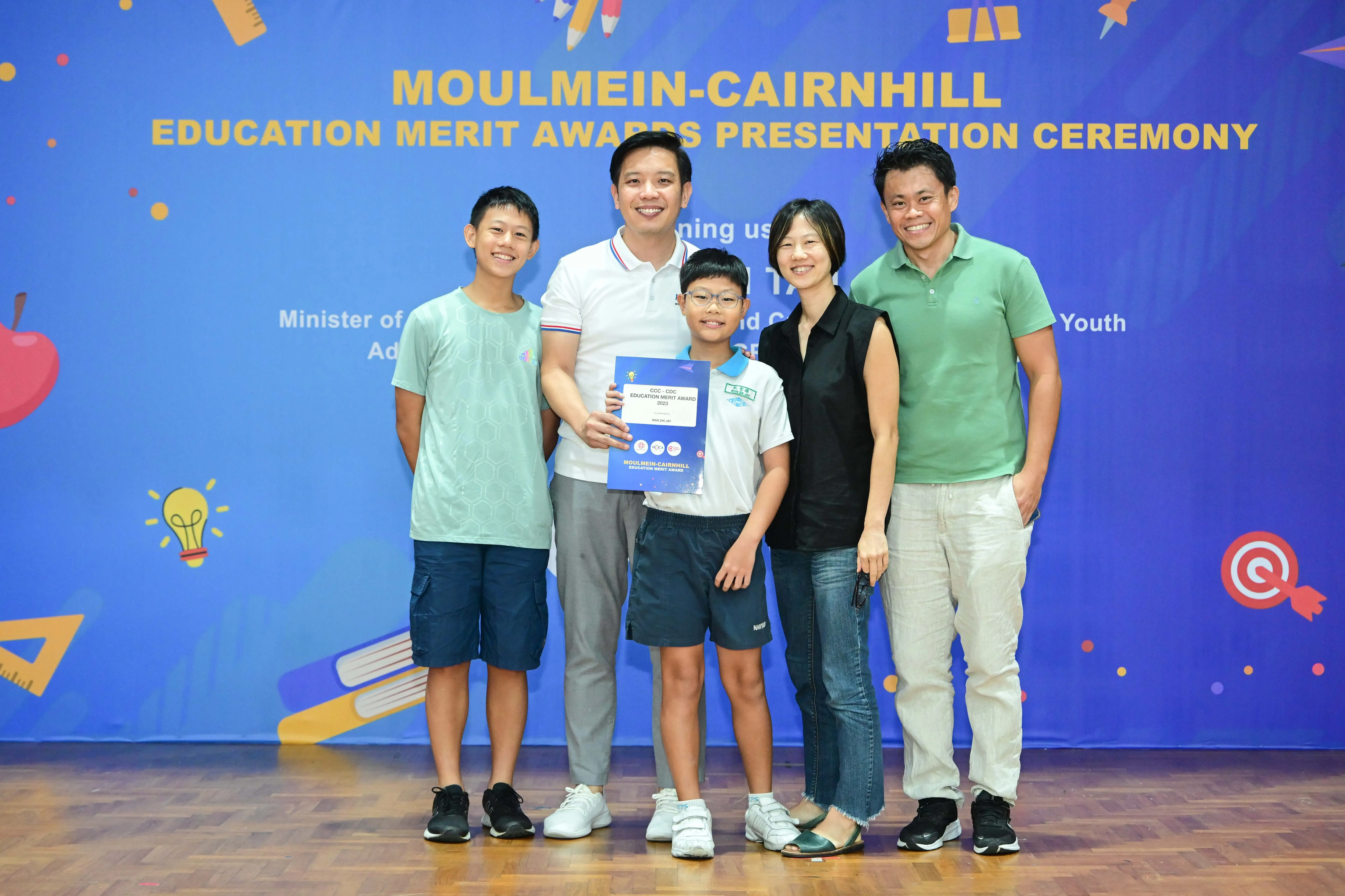 Five people pose smiling before a Moulmein-Cairnhill Education Merit Awards banner, with a child holding a certificate.