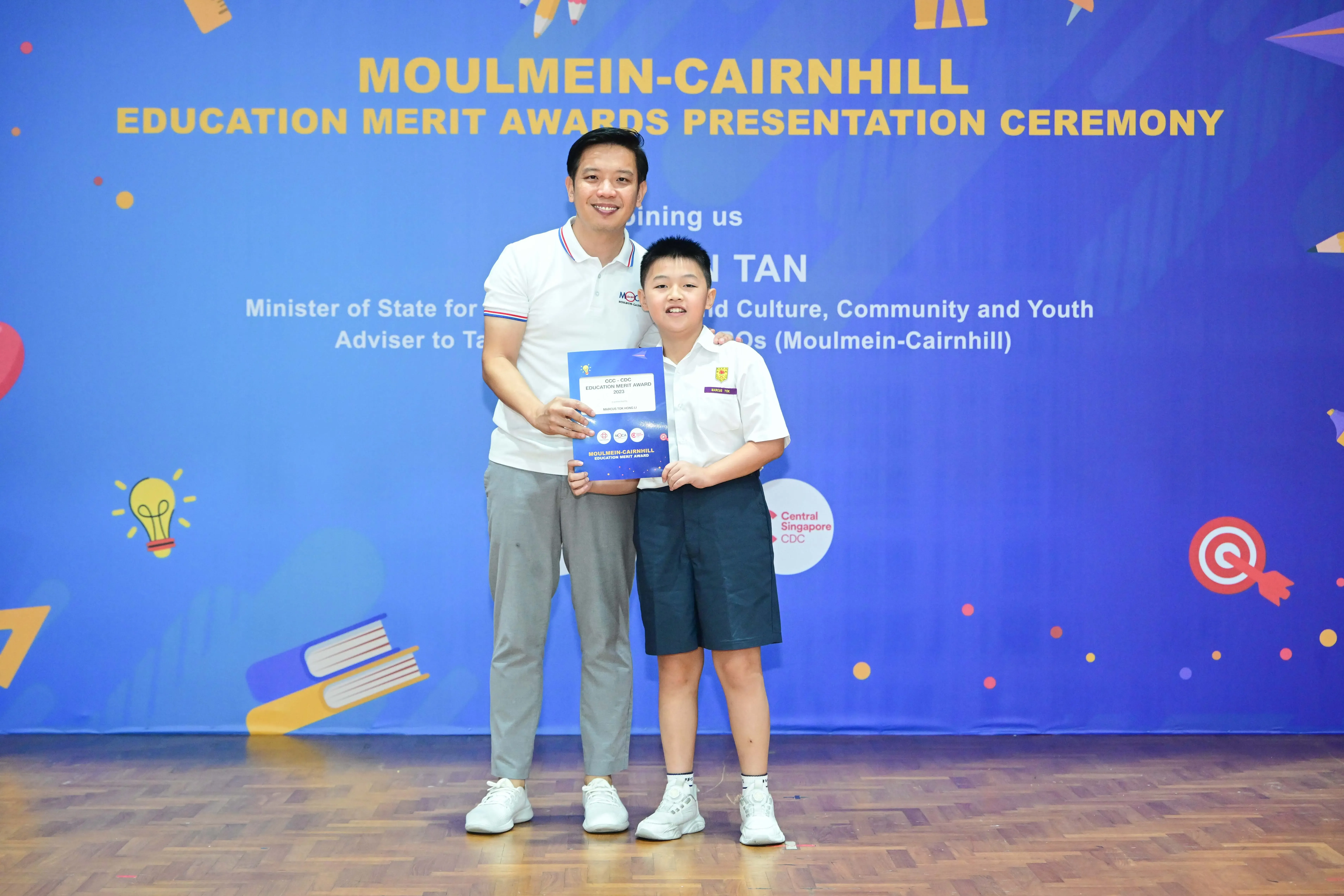An adult and a young boy hold an award certificate at the Moulmein-Cairnhill Education Merit Awards Presentation Ceremony.