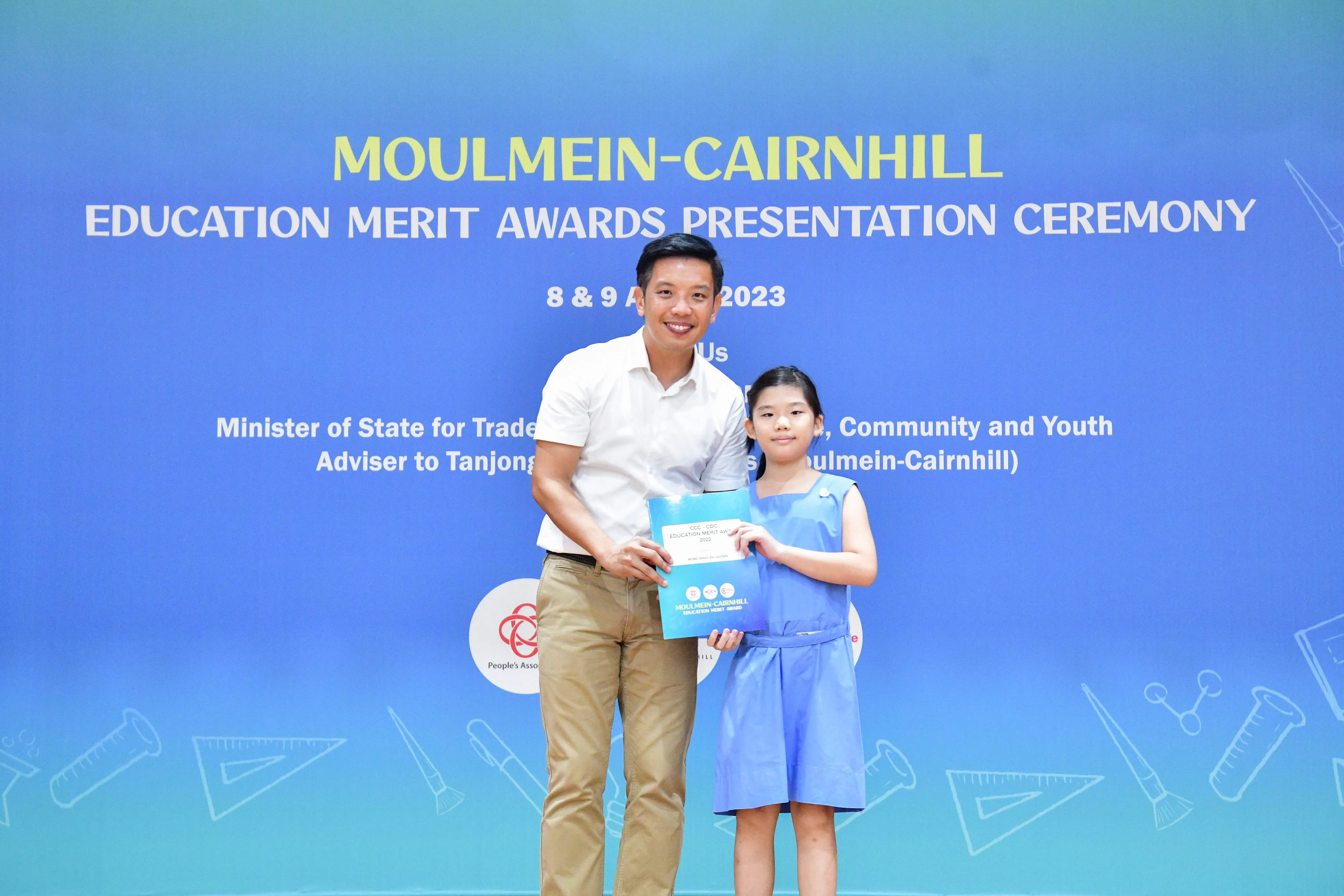 A man and a young girl hold an award folder in front of a Moulmein-Cairnhill Education Merit Awards backdrop.
