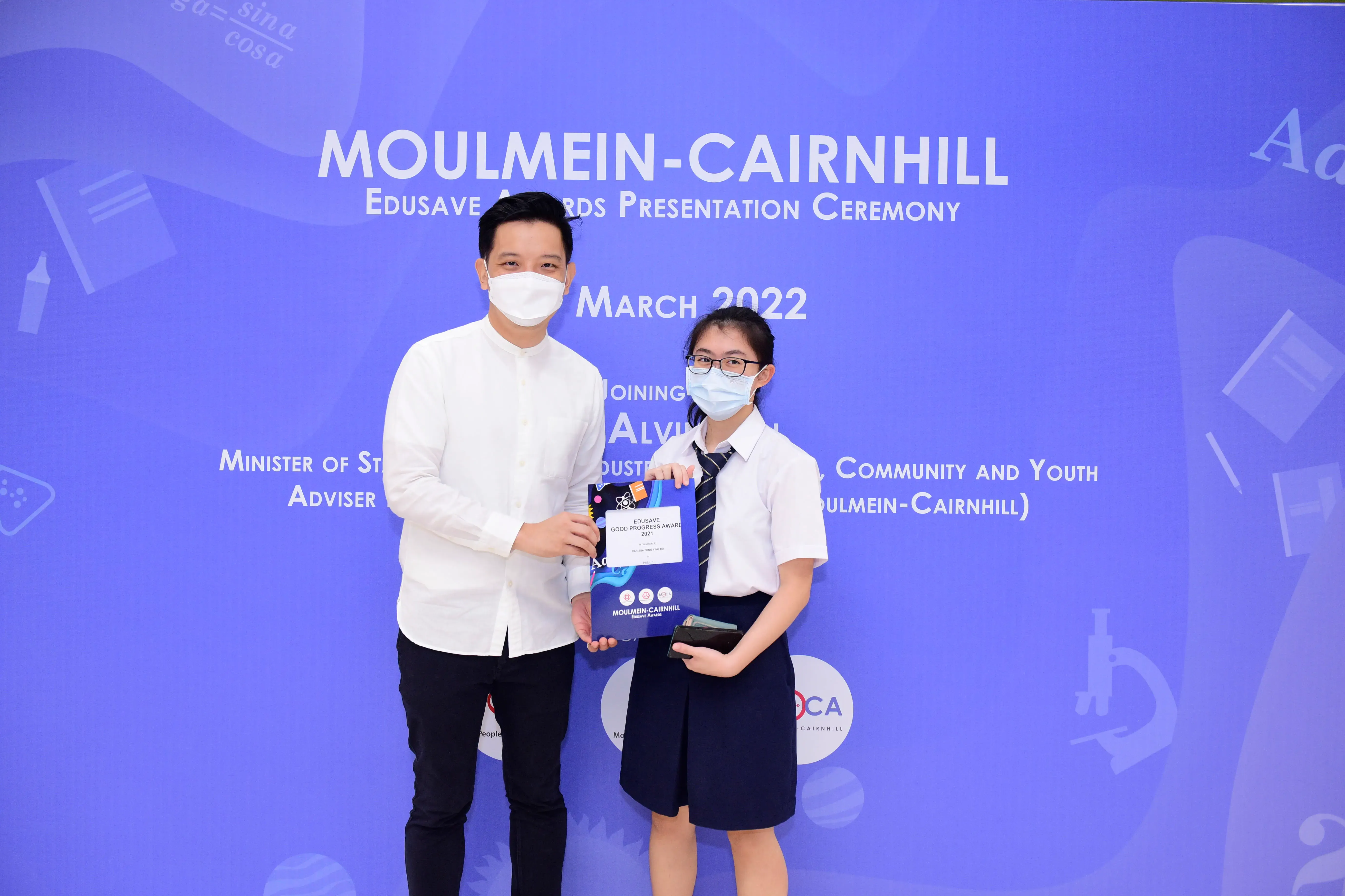 Two masked people hold an Edusave Good Progress Award at the Moulmein-Cairnhill Awards Ceremony, March 2022.