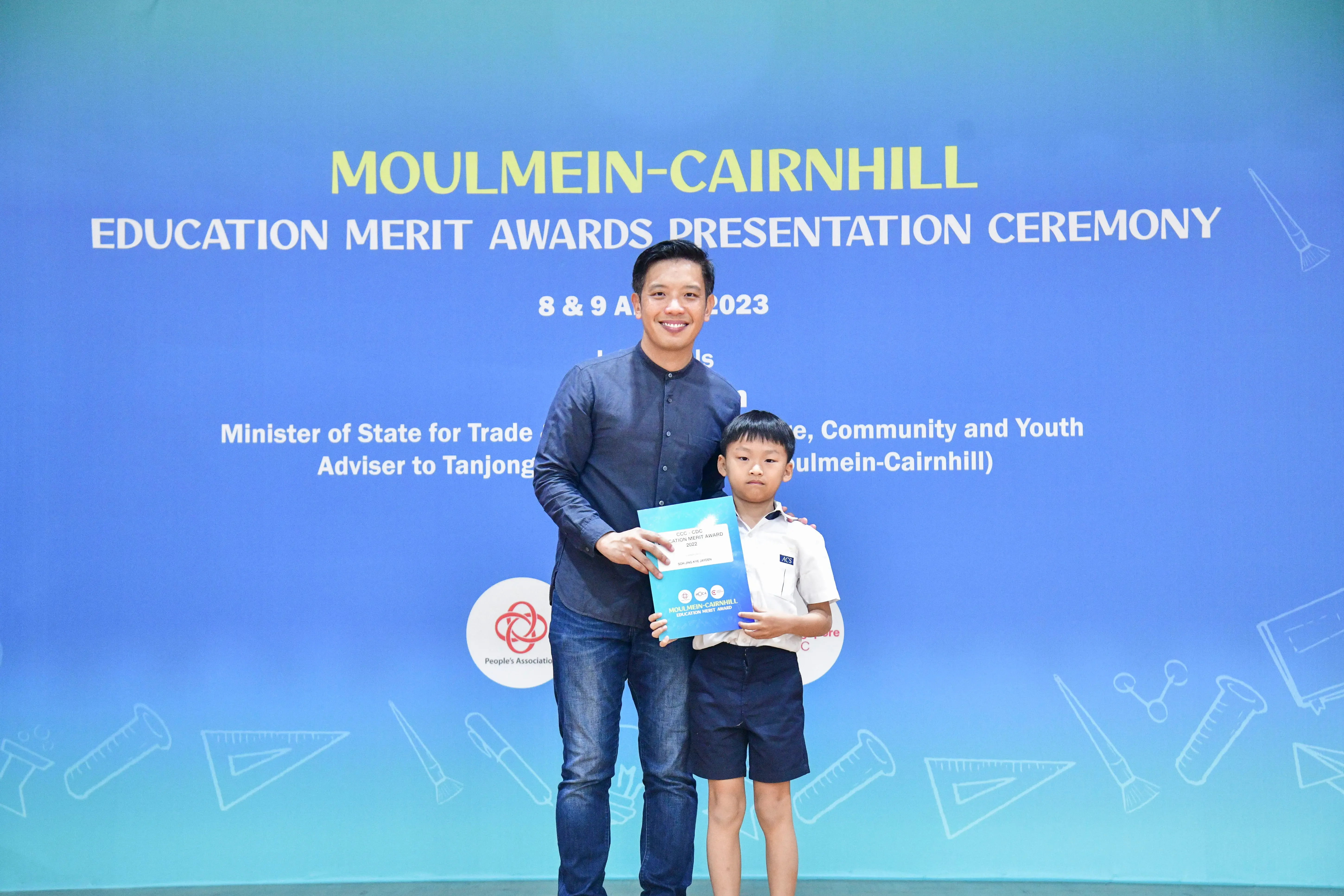 A man and a young boy hold a certificate in front of a blue Moulmein-Cairnhill Education Merit Awards banner.