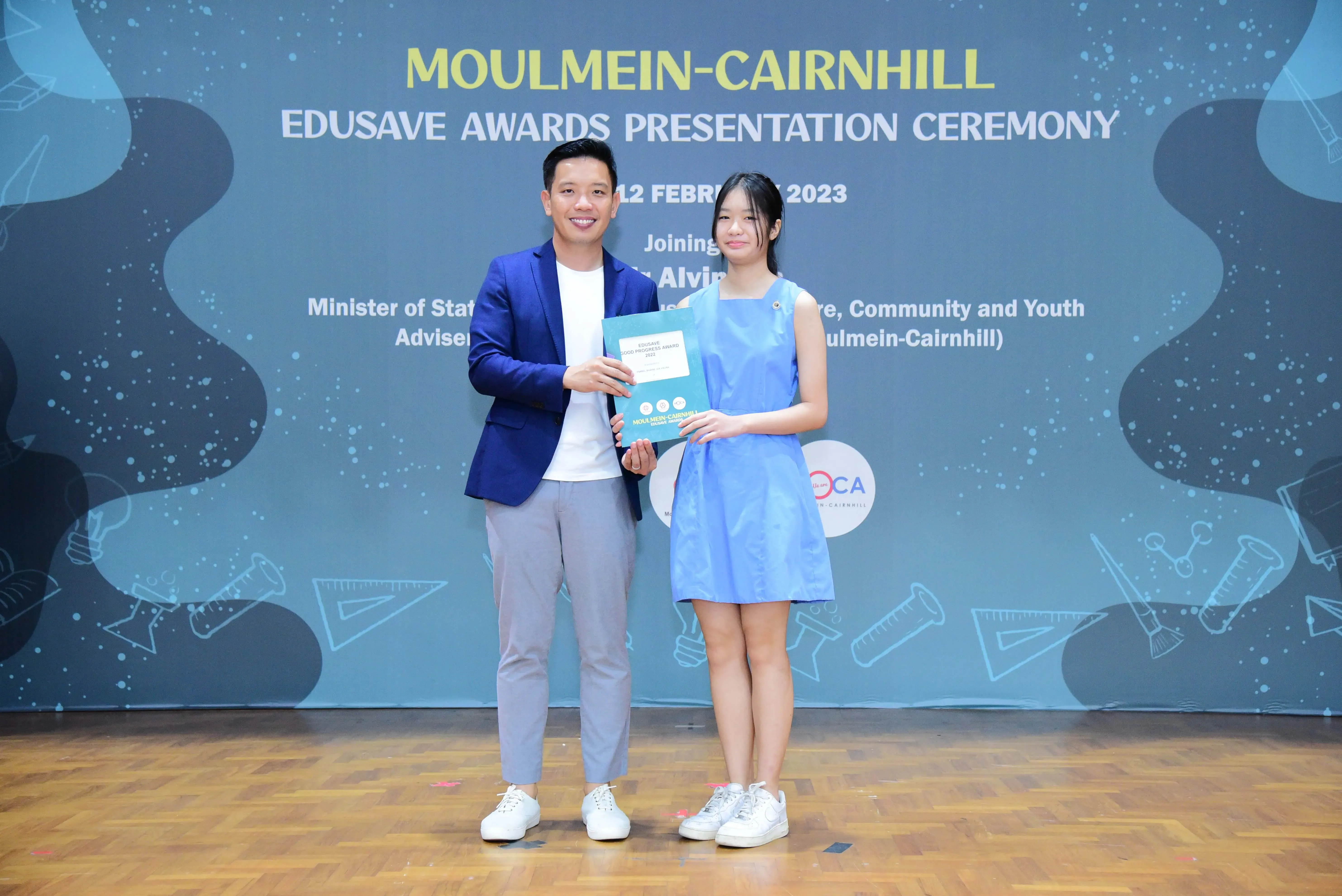 Two people hold a certificate together on stage at the Moulmein-Cairnhill Edusave Awards Presentation Ceremony 2023.