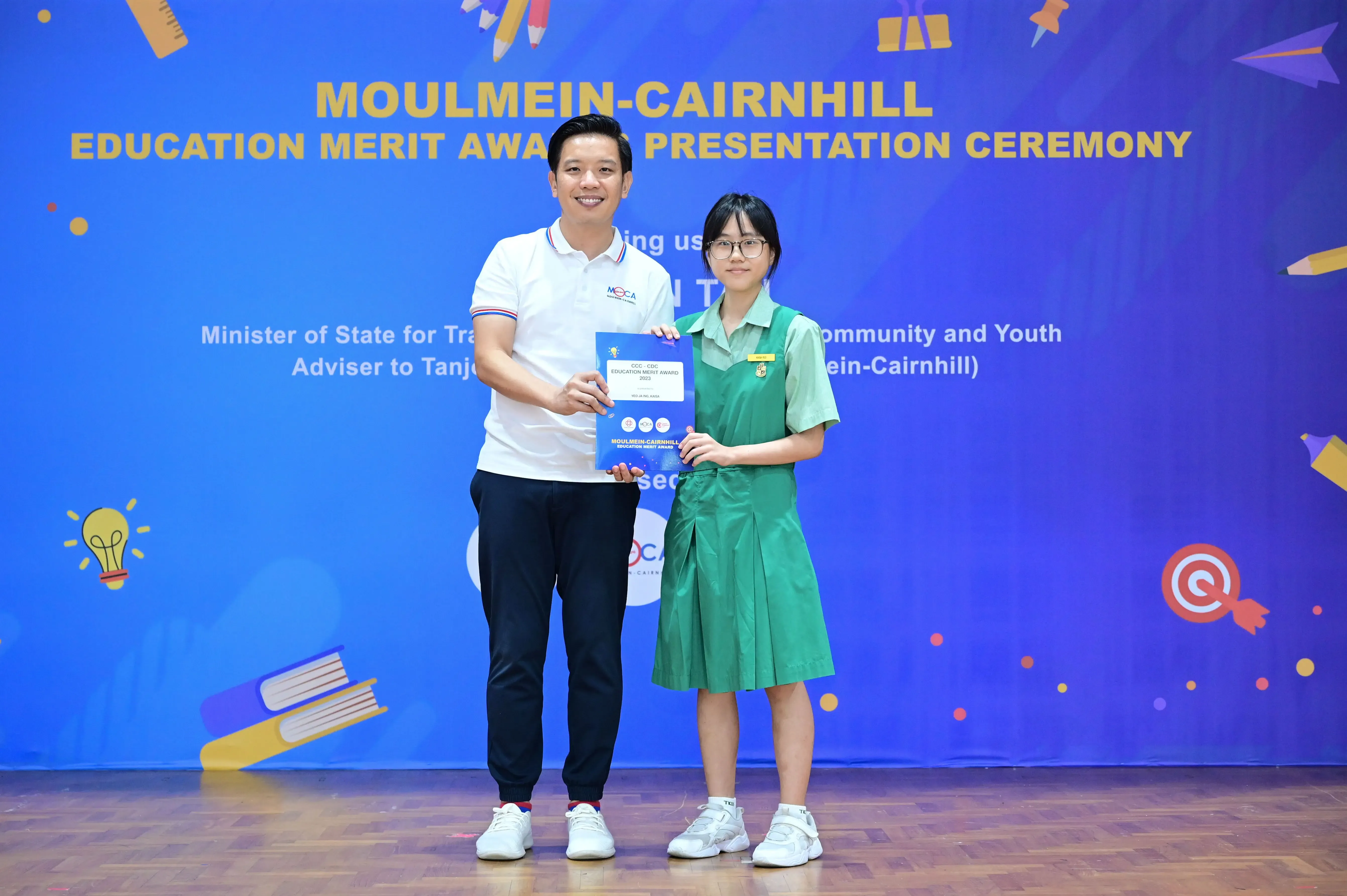 A man and a young girl in a green school uniform hold an award certificate at the Moulmein-Cairnhill Education Merit Award ceremony.