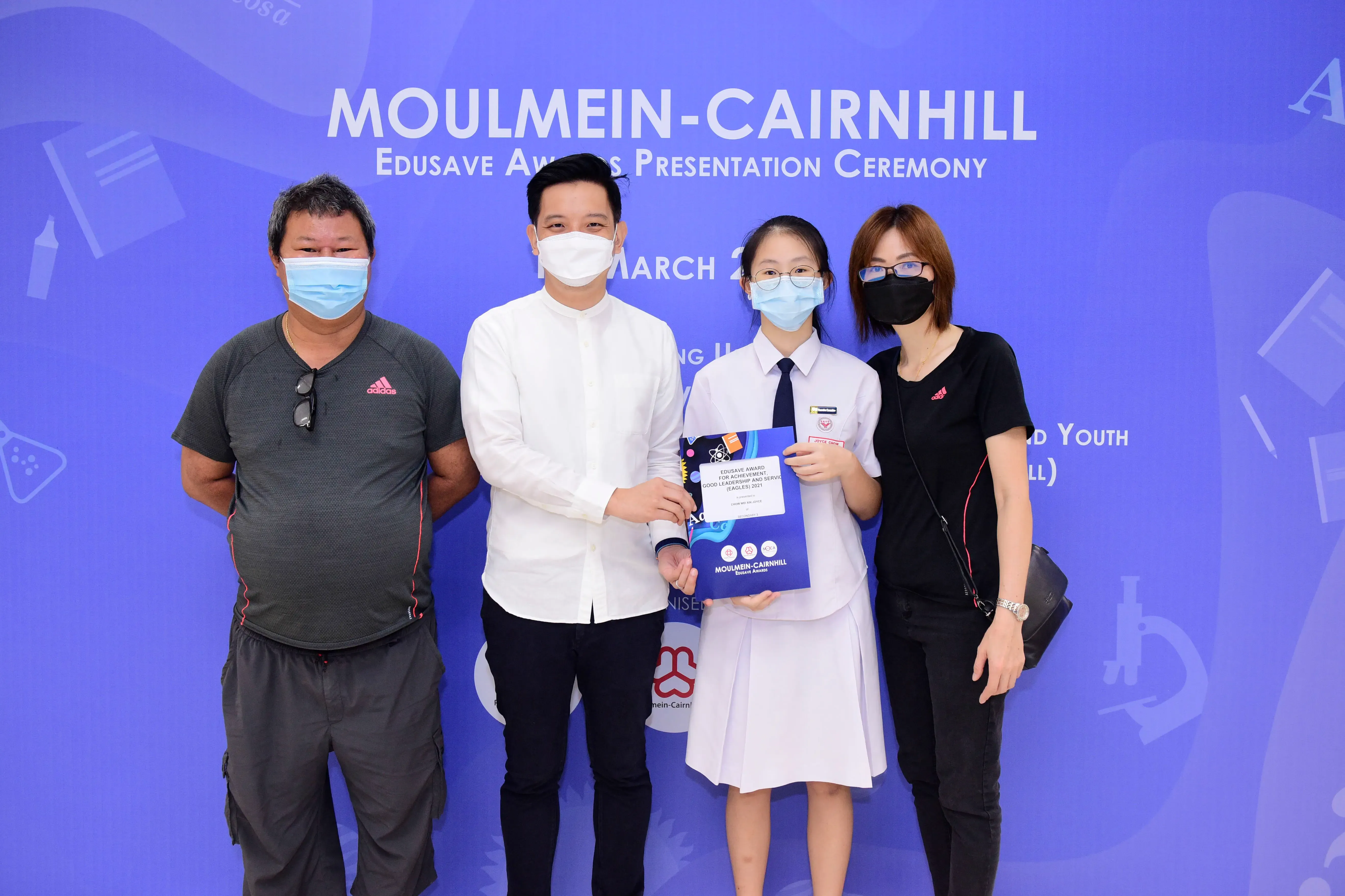 Four masked people pose at Moulmein-Cairnhill Edusave Awards Presentation Ceremony, with a student holding an award certificate.