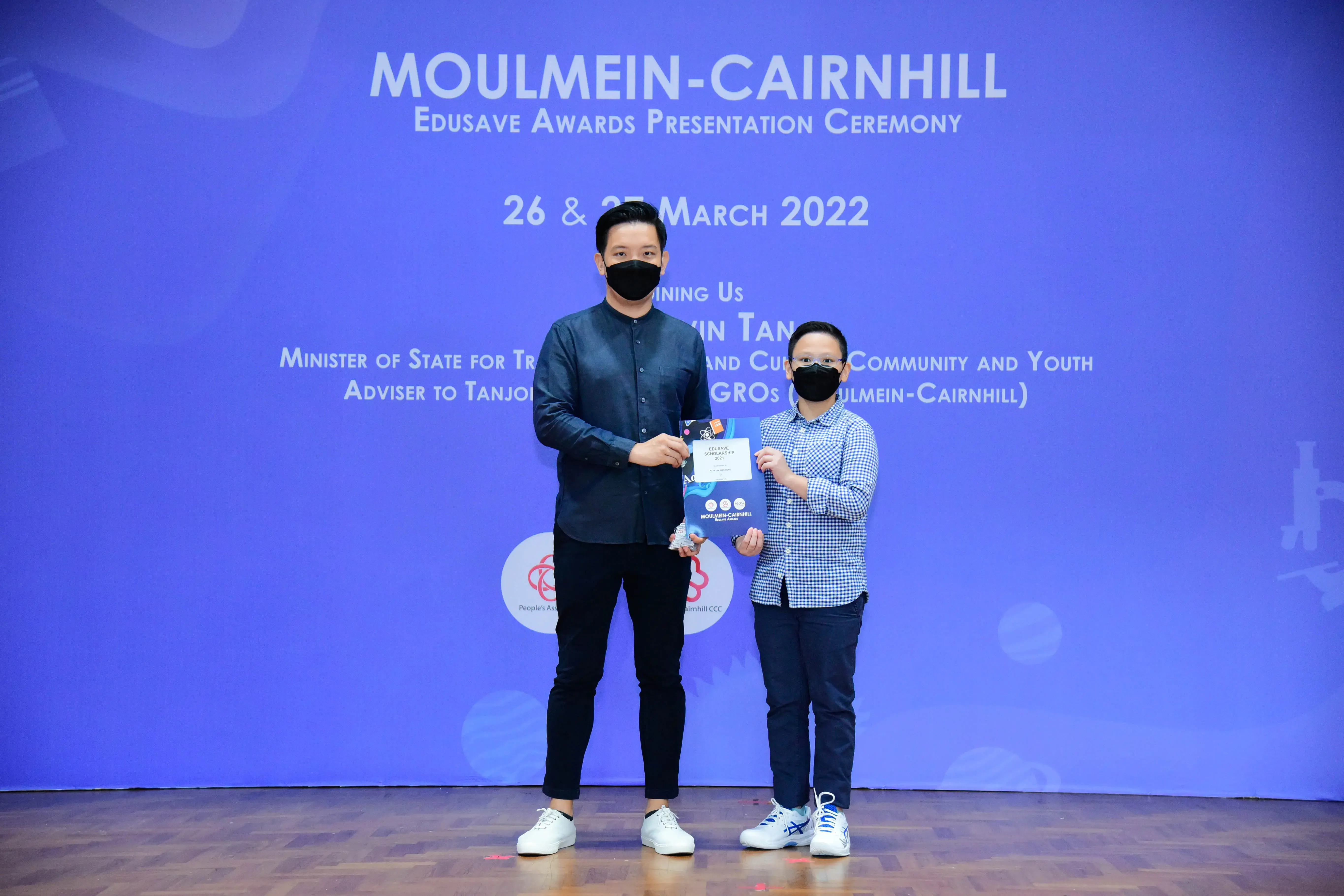 Two masked men stand on a stage holding an award at the Moulmein-Cairnhill Edusave Awards Ceremony, March 2022.