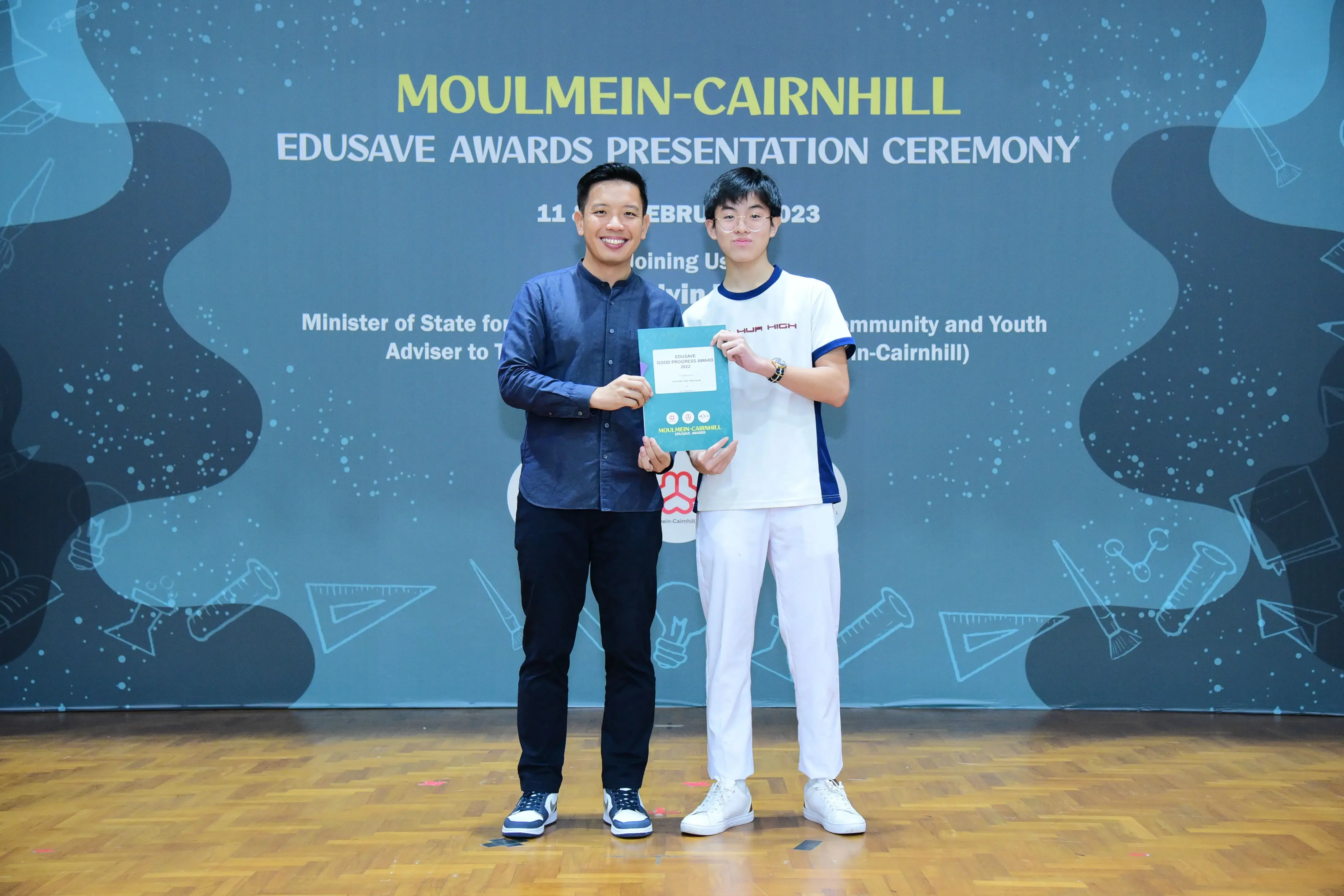 Two men hold a certificate together on stage at the Moulmein-Cairnhill Edusave Awards Presentation Ceremony 2023.