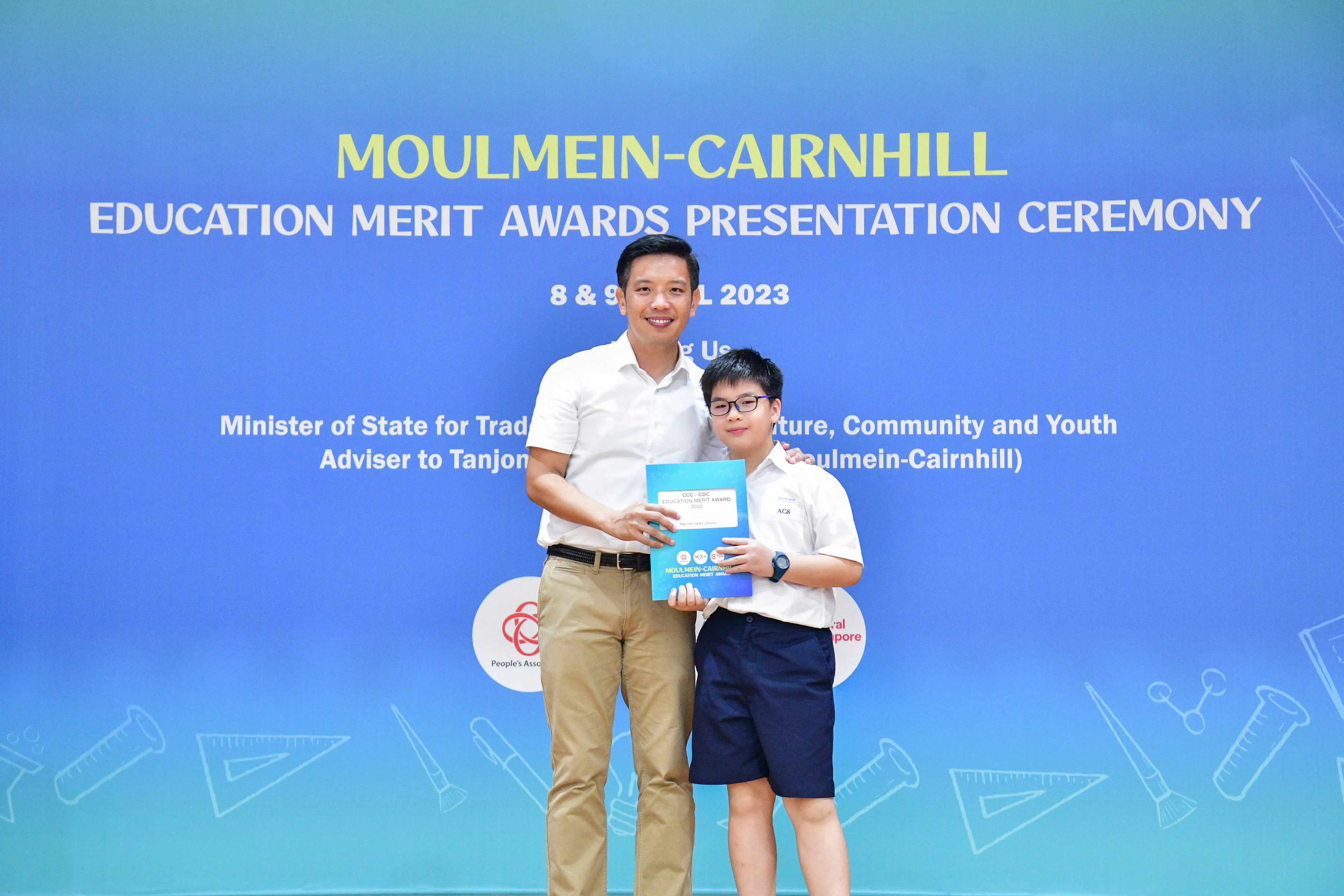 A man and a young boy in school uniform hold a blue award folder at the Moulmein-Cairnhill Education Merit Awards 2023.