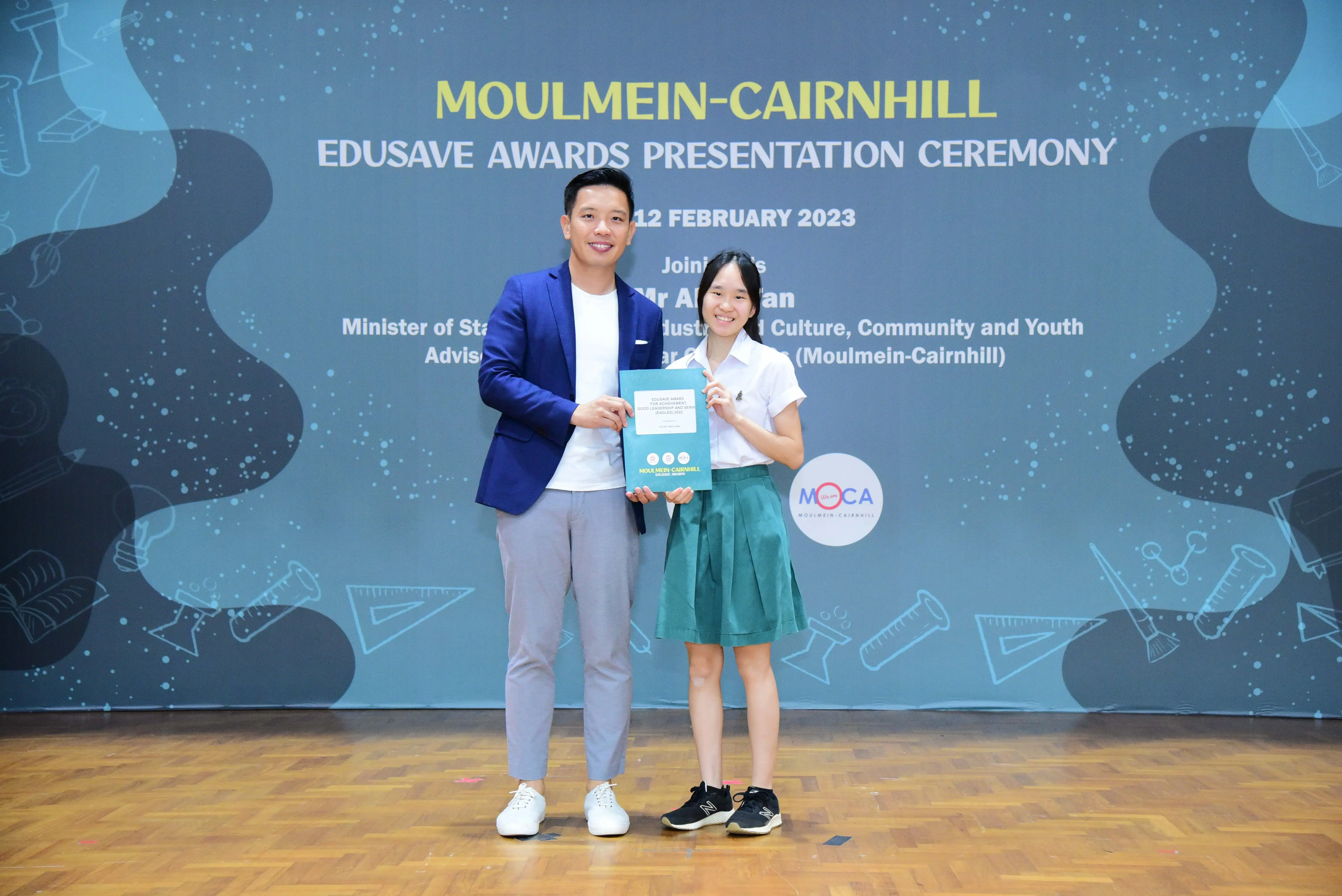 A man in a blue blazer and a girl in school uniform hold an award at the Moulmein-Cairnhill Edusave Awards Ceremony 2023.