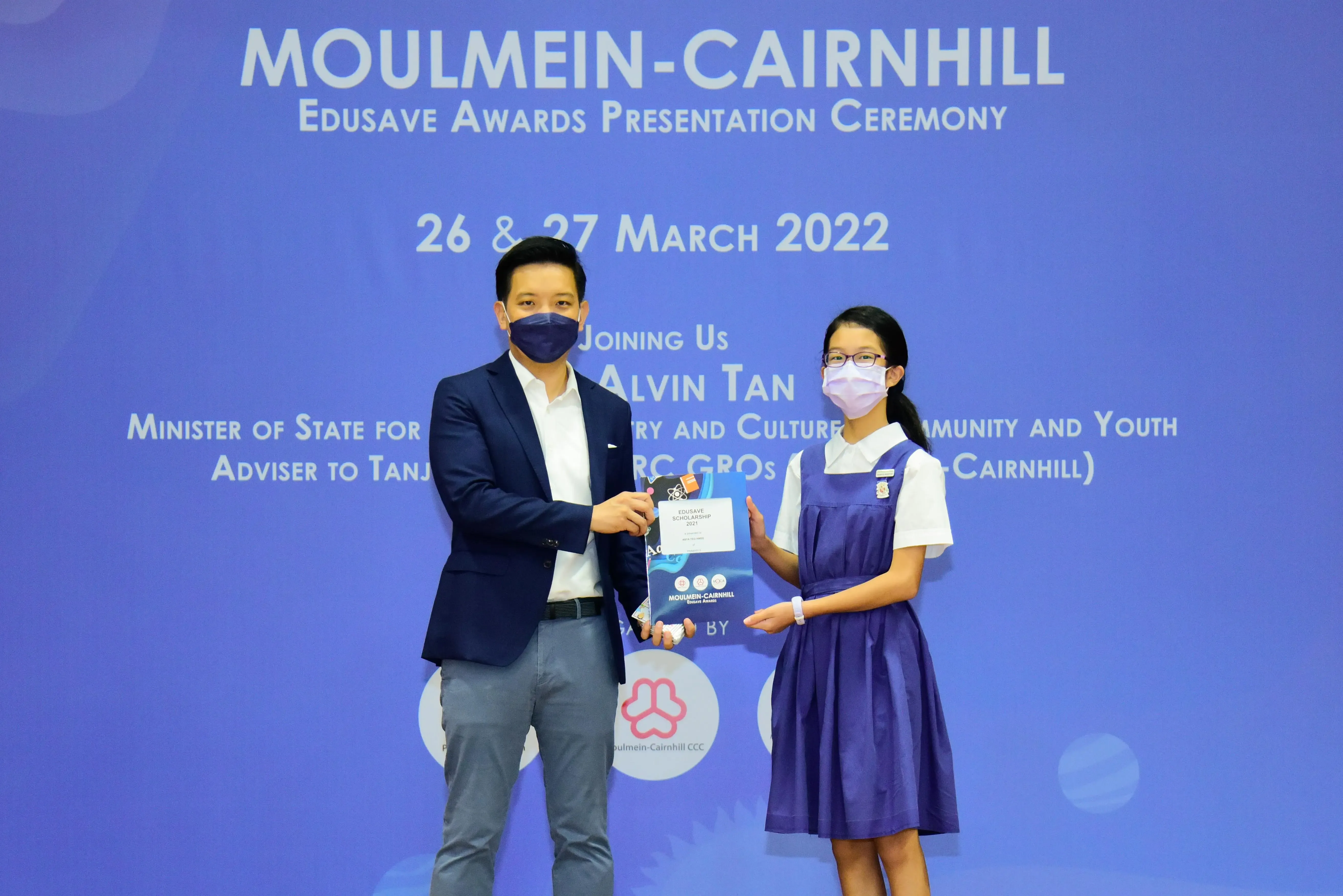 A man in a suit and a girl in school uniform hold an award certificate at the Moulmein-Cairnhill Edusave ceremony.
