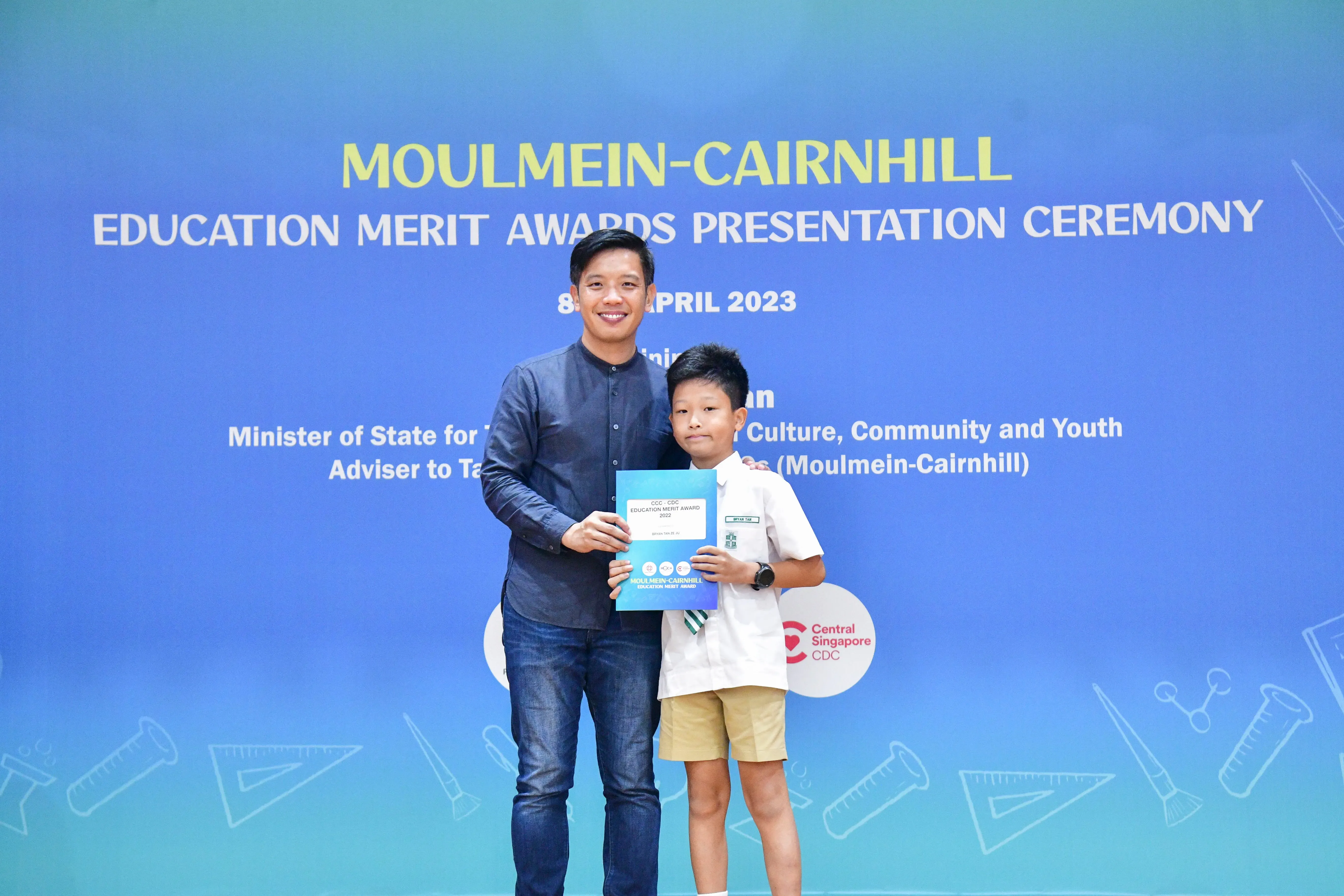 A man and a young boy hold a certificate in front of a blue Moulmein-Cairnhill Education Merit Awards banner.