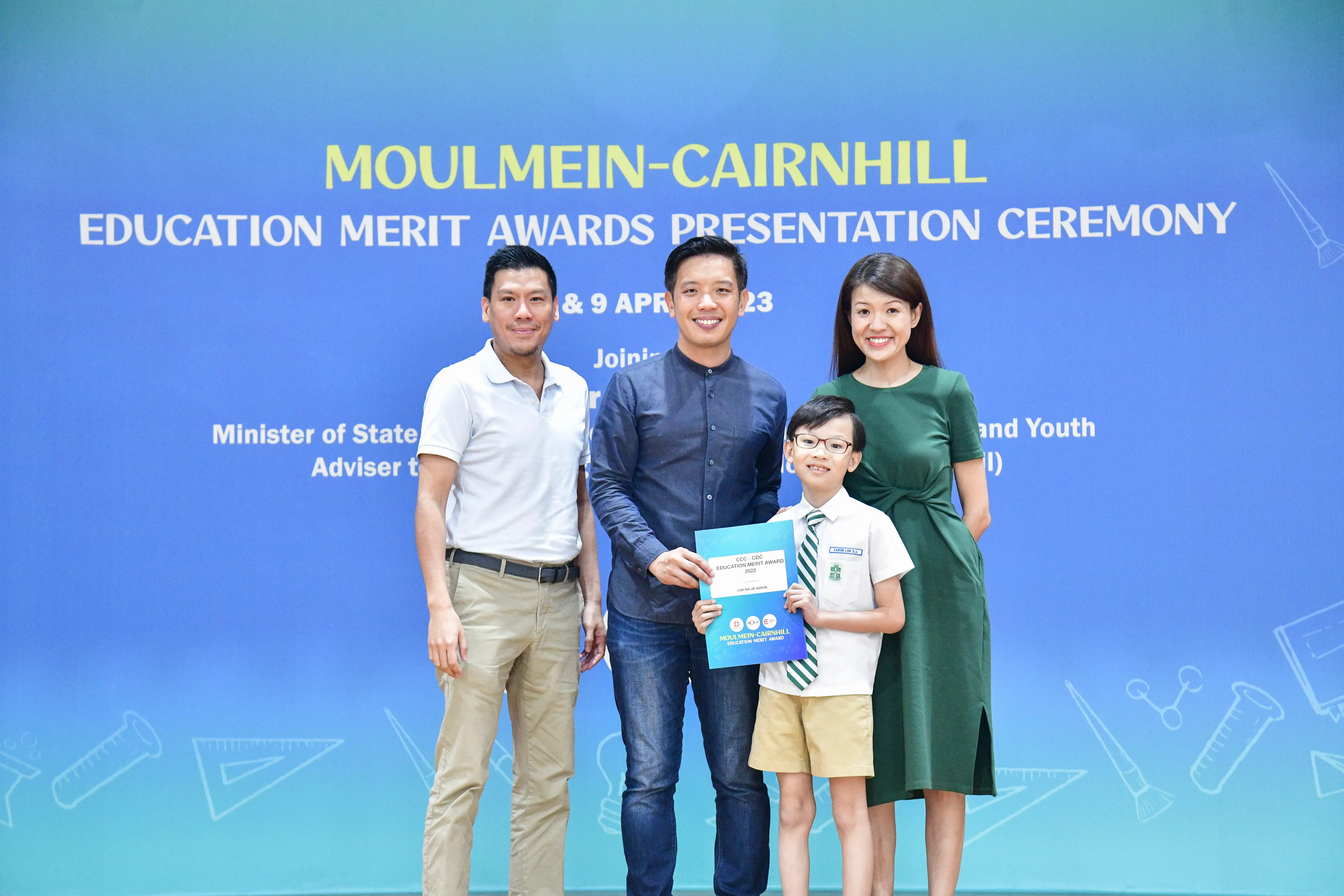 Three adults and a young boy in school uniform pose at Moulmein-Cairnhill Education Merit Awards Ceremony holding a certificate.
