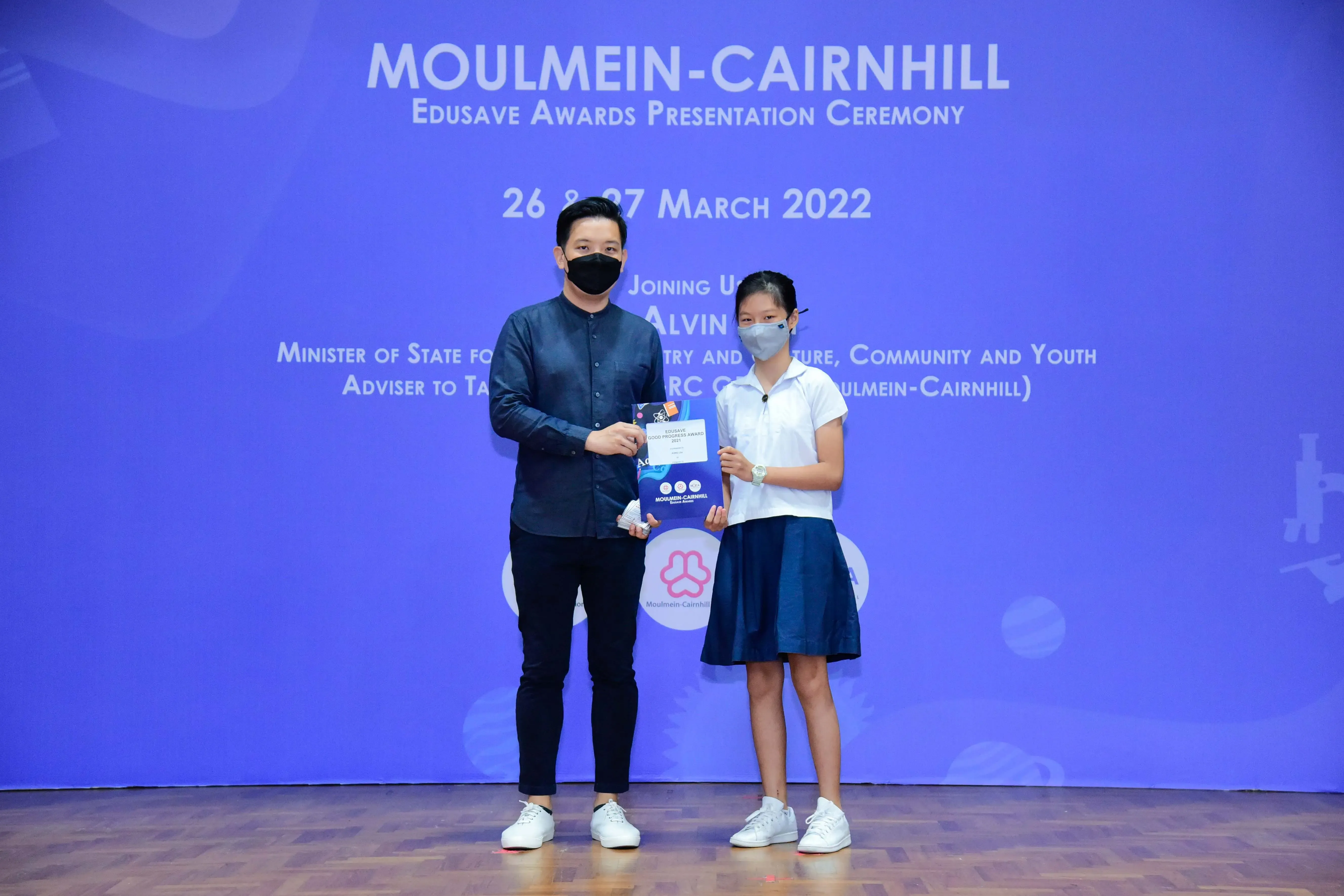 A masked man and a young girl hold a certificate on stage at the Moulmein-Cairnhill Edusave Awards Ceremony, March 2022.