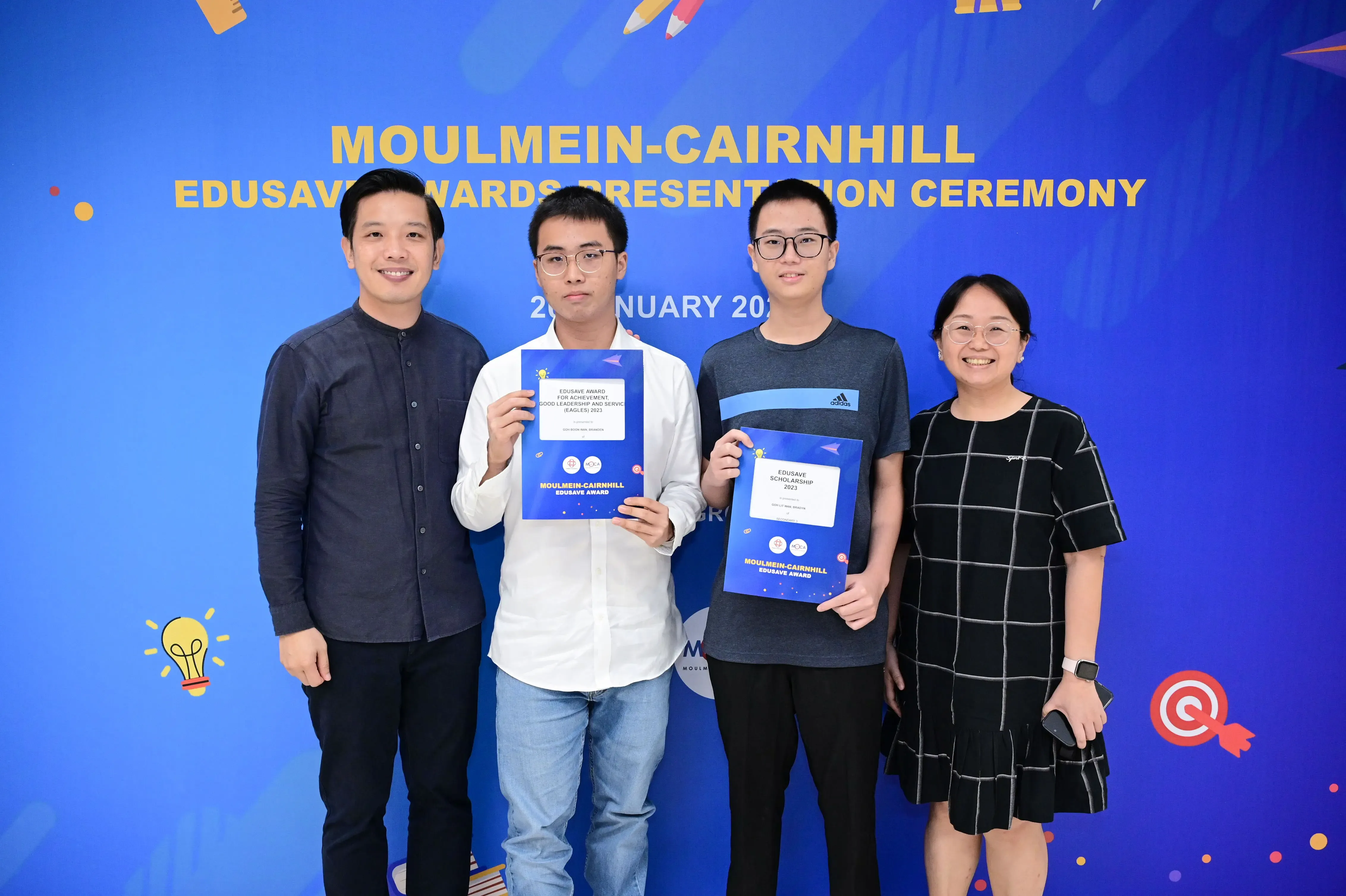 Four people pose in front of a Moulmein-Cairnhill Edusave Awards Presentation Ceremony banner, two holding award certificates.