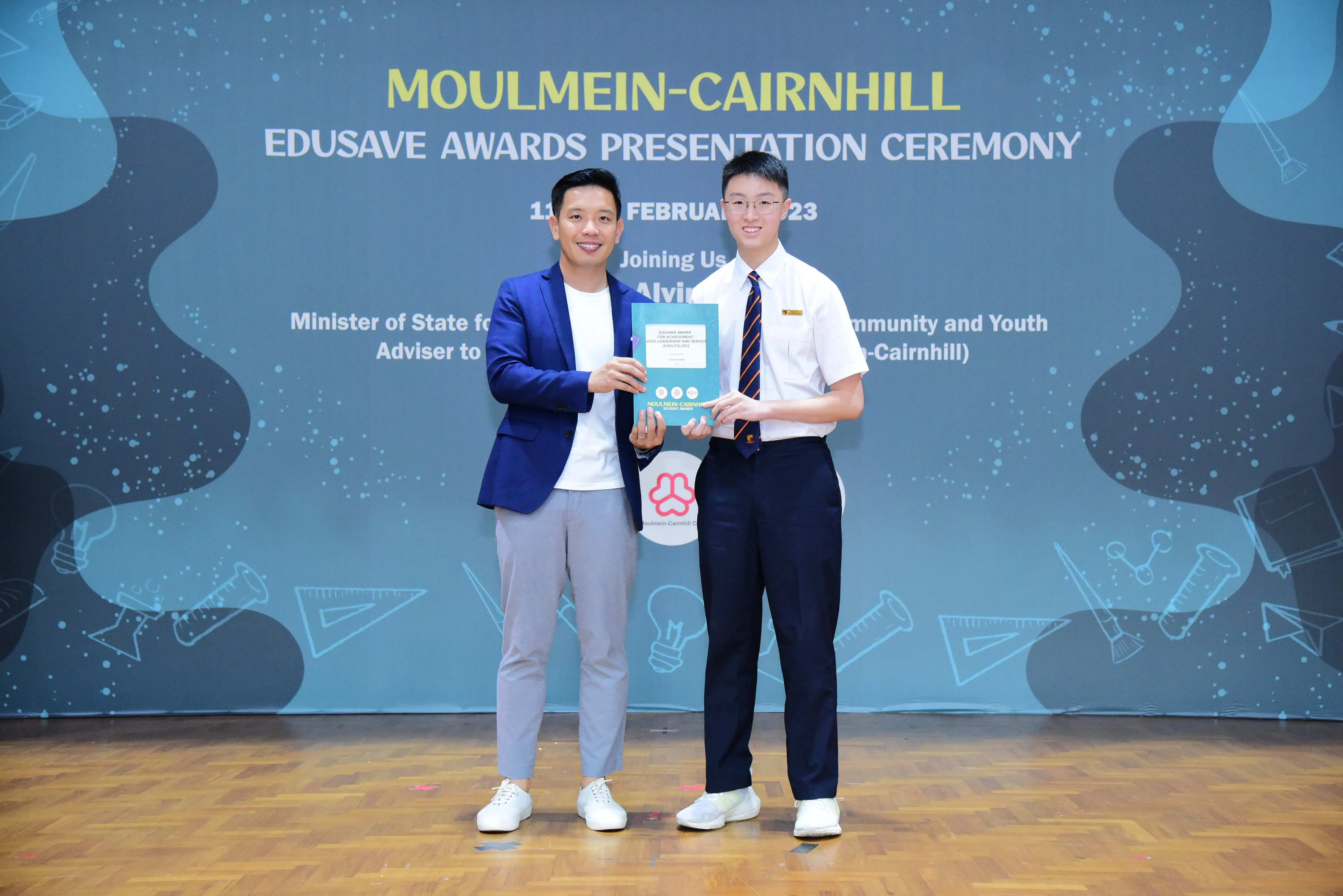 Two men hold a certificate together on stage at the Moulmein-Cairnhill Edusave Awards Presentation Ceremony 2023.
