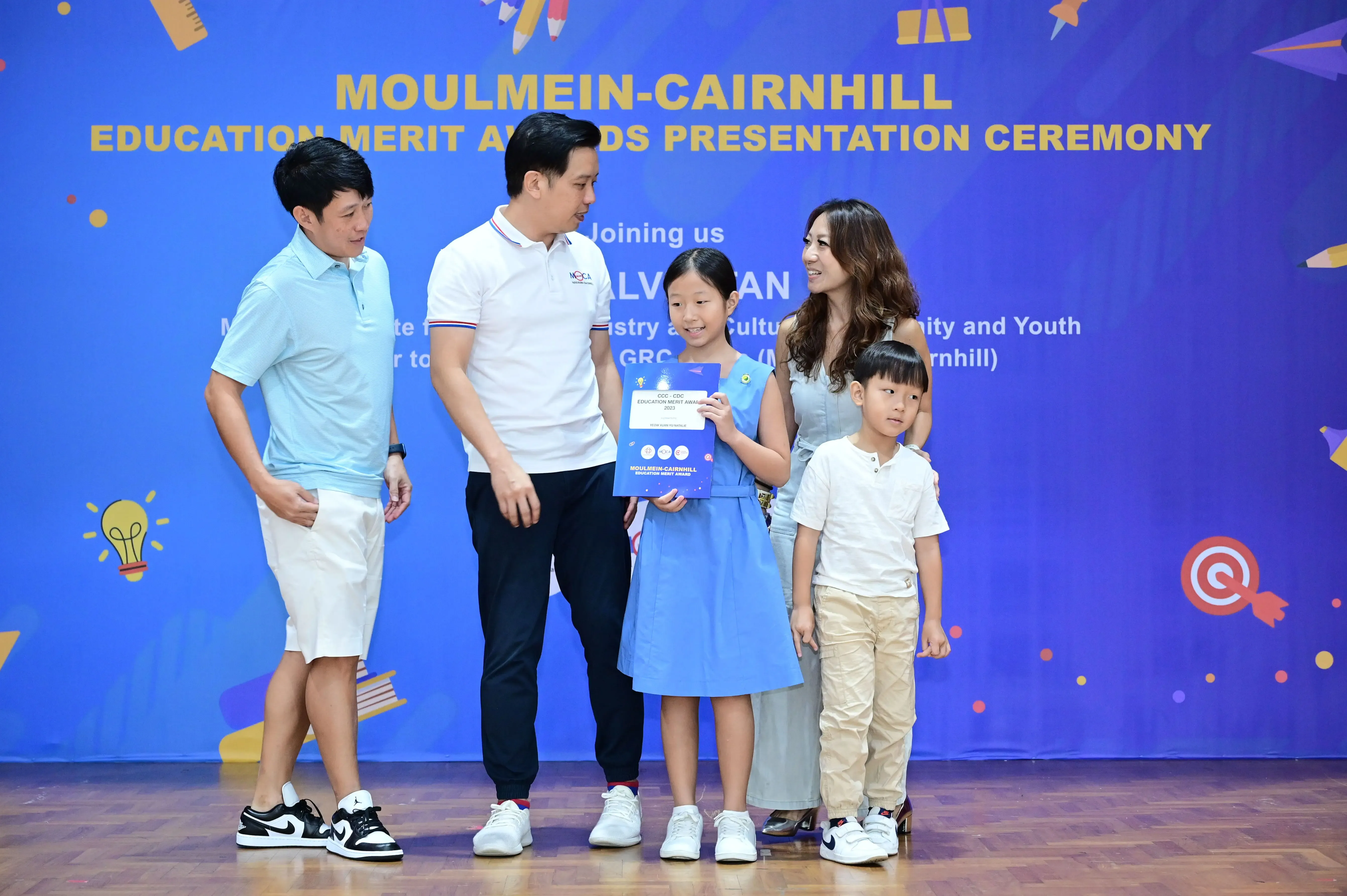A girl holds a certificate at the Moulmein-Cairnhill Education Merit Awards ceremony, standing with two men, a woman, and a boy.