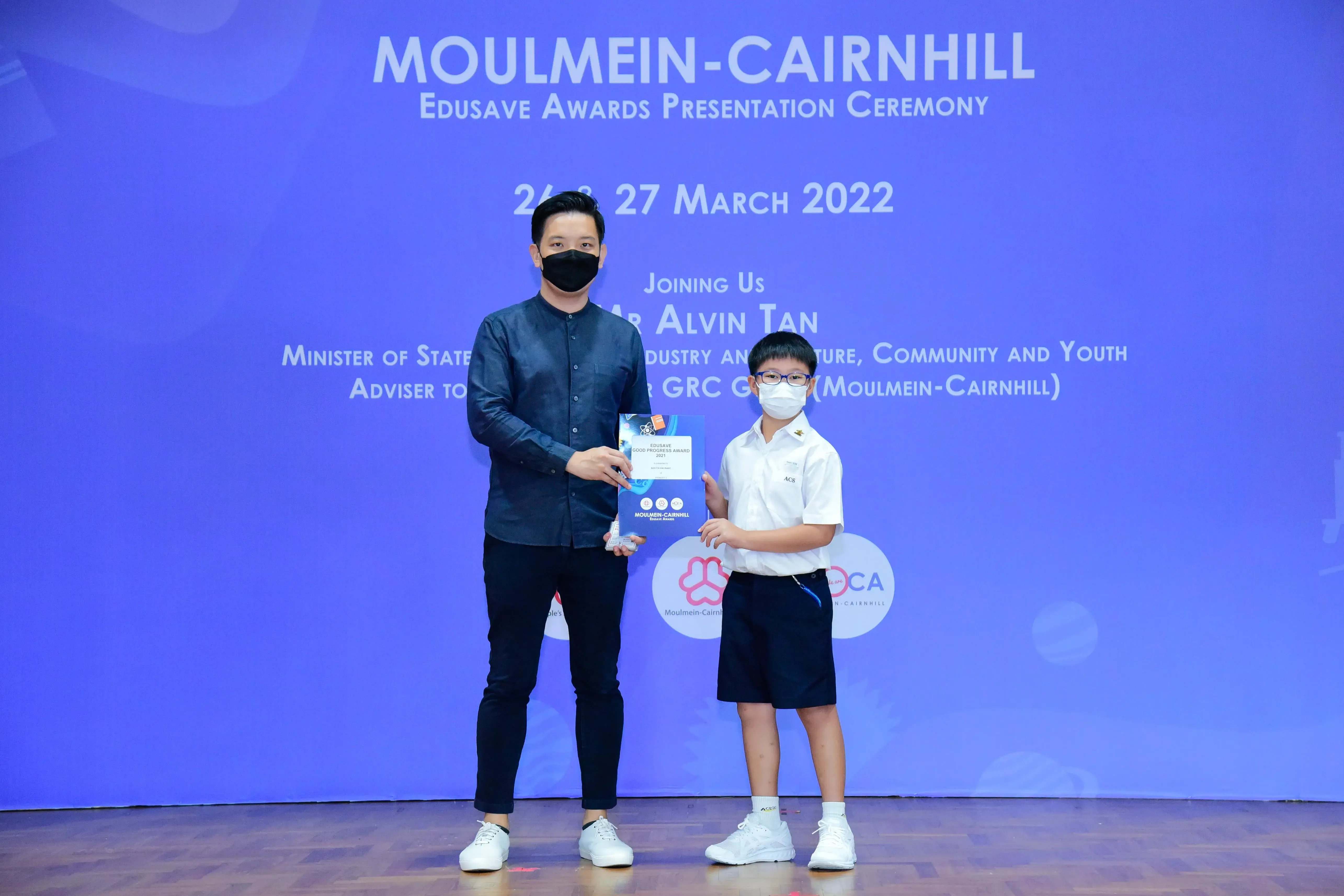 An adult and a young student in masks hold a certificate on stage at the Moulmein-Cairnhill Edusave Awards 2022.