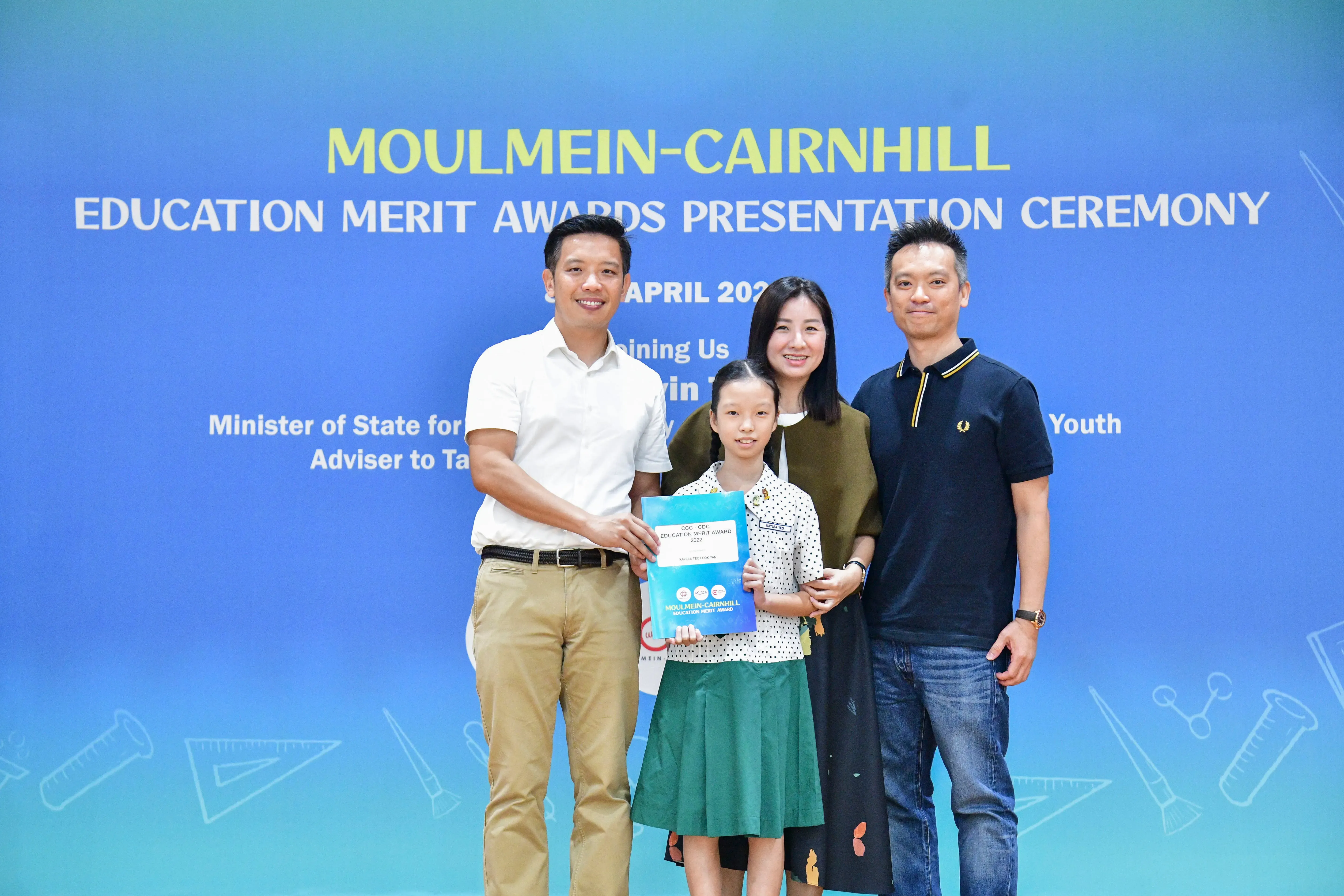 A young girl holds an award certificate with three adults at the Moulmein-Cairnhill Education Merit Awards Ceremony.