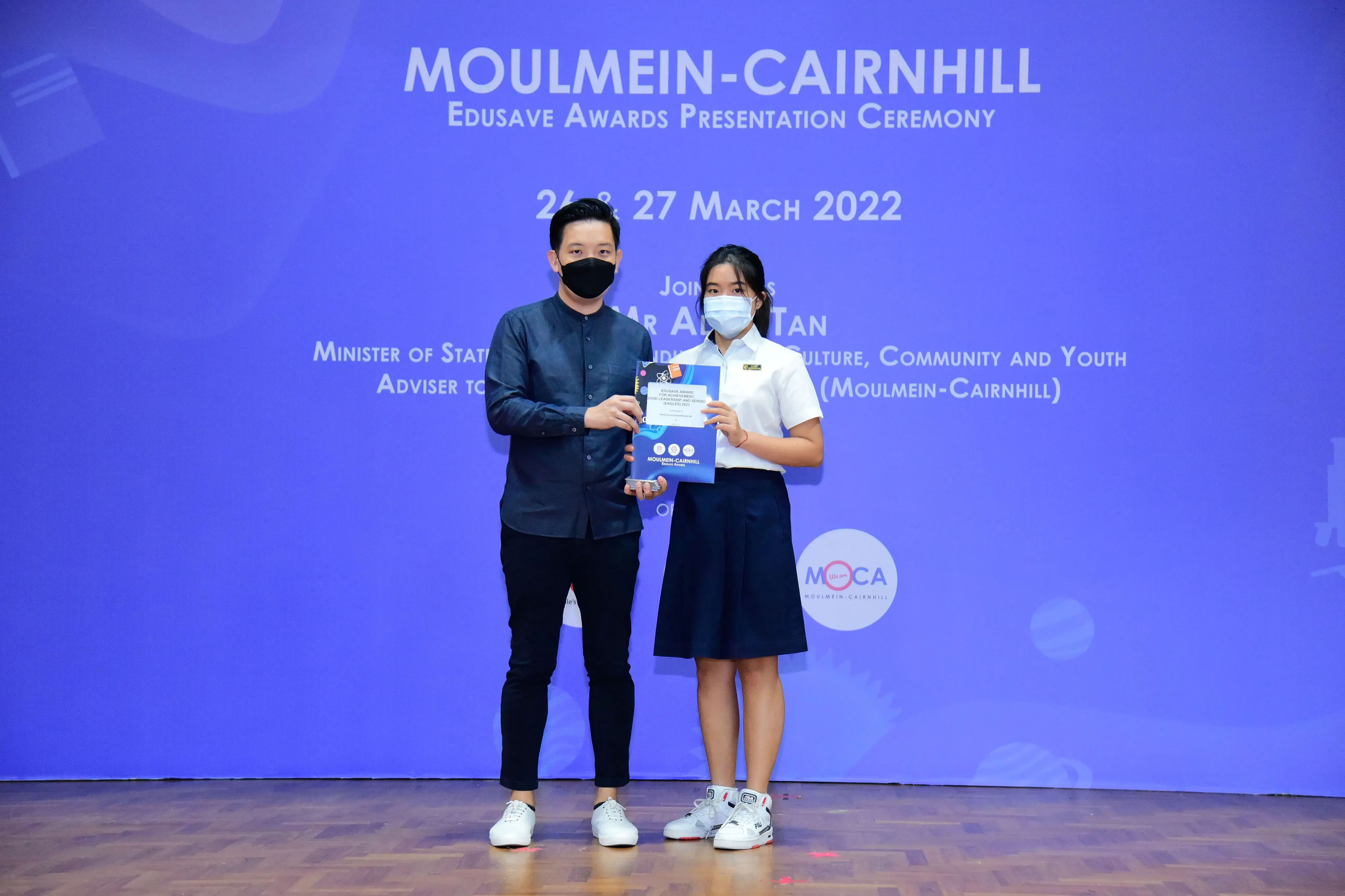 Two masked individuals hold an award folder on stage at the Moulmein-Cairnhill Edusave Awards Ceremony, March 2022.
