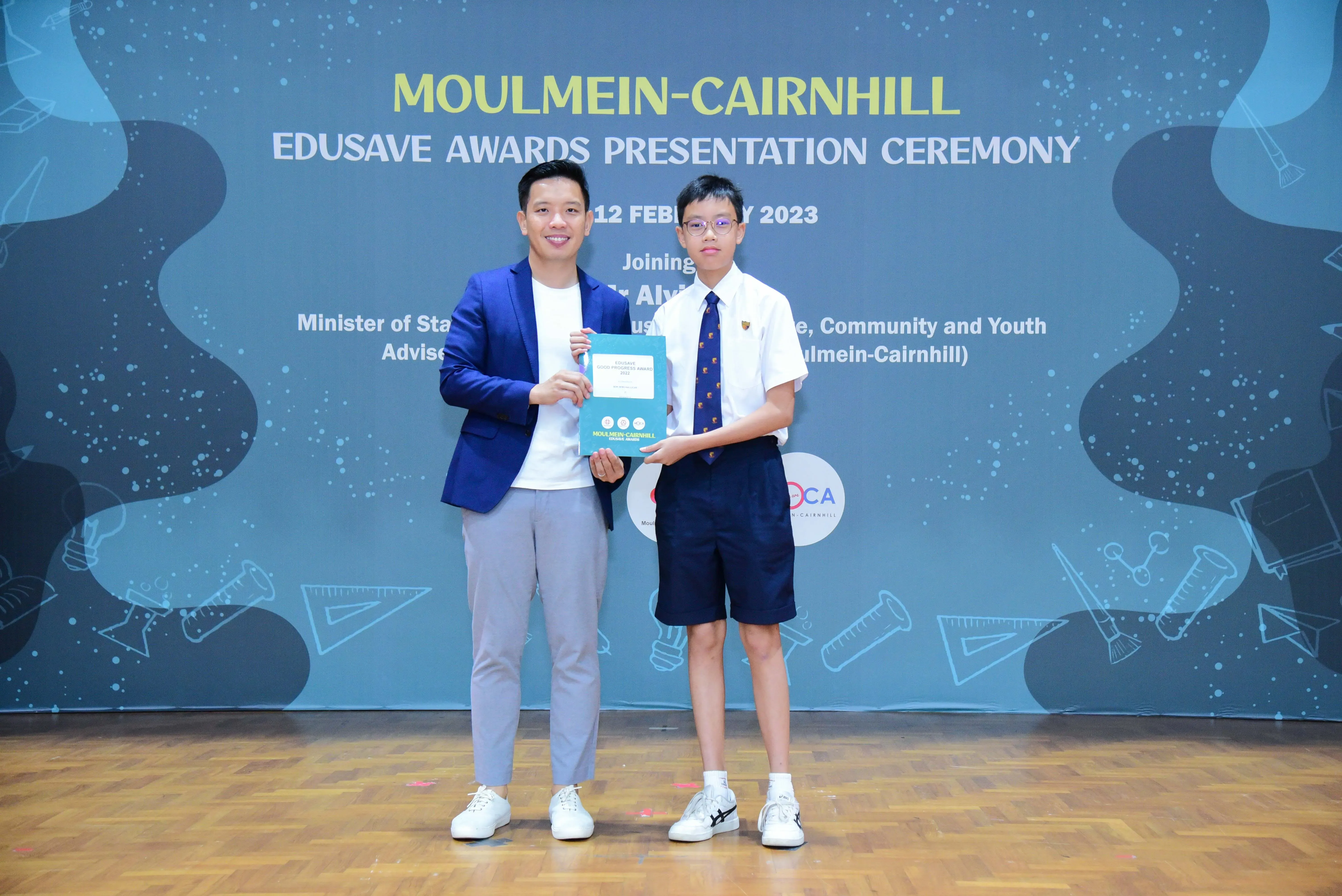 A man in a blue blazer and a young student in uniform hold a certificate at the Moulmein-Cairnhill Edusave Awards Ceremony 2023.