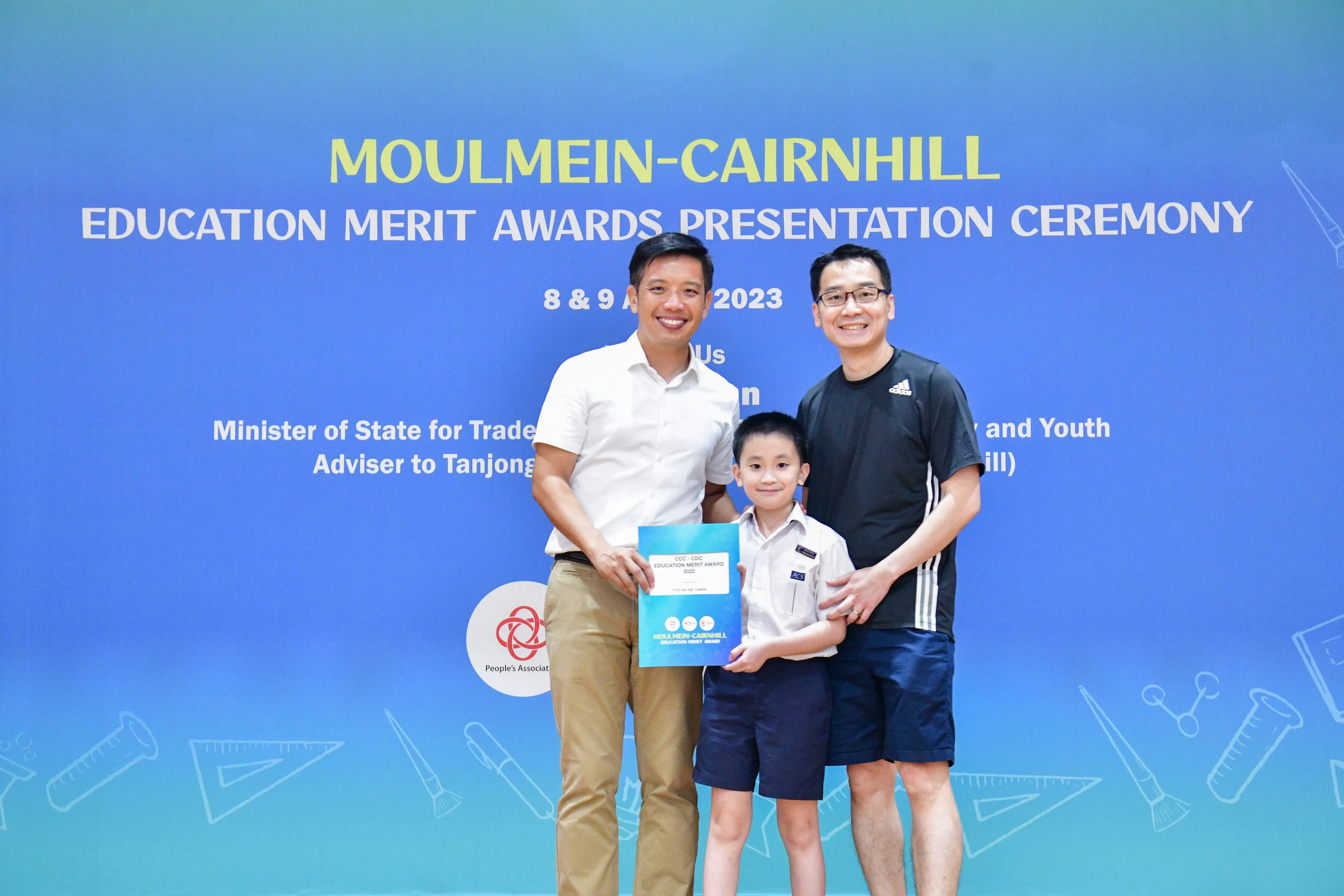 Two men and a young boy holding an award certificate pose before a Moulmein-Cairnhill Education Merit Awards banner.