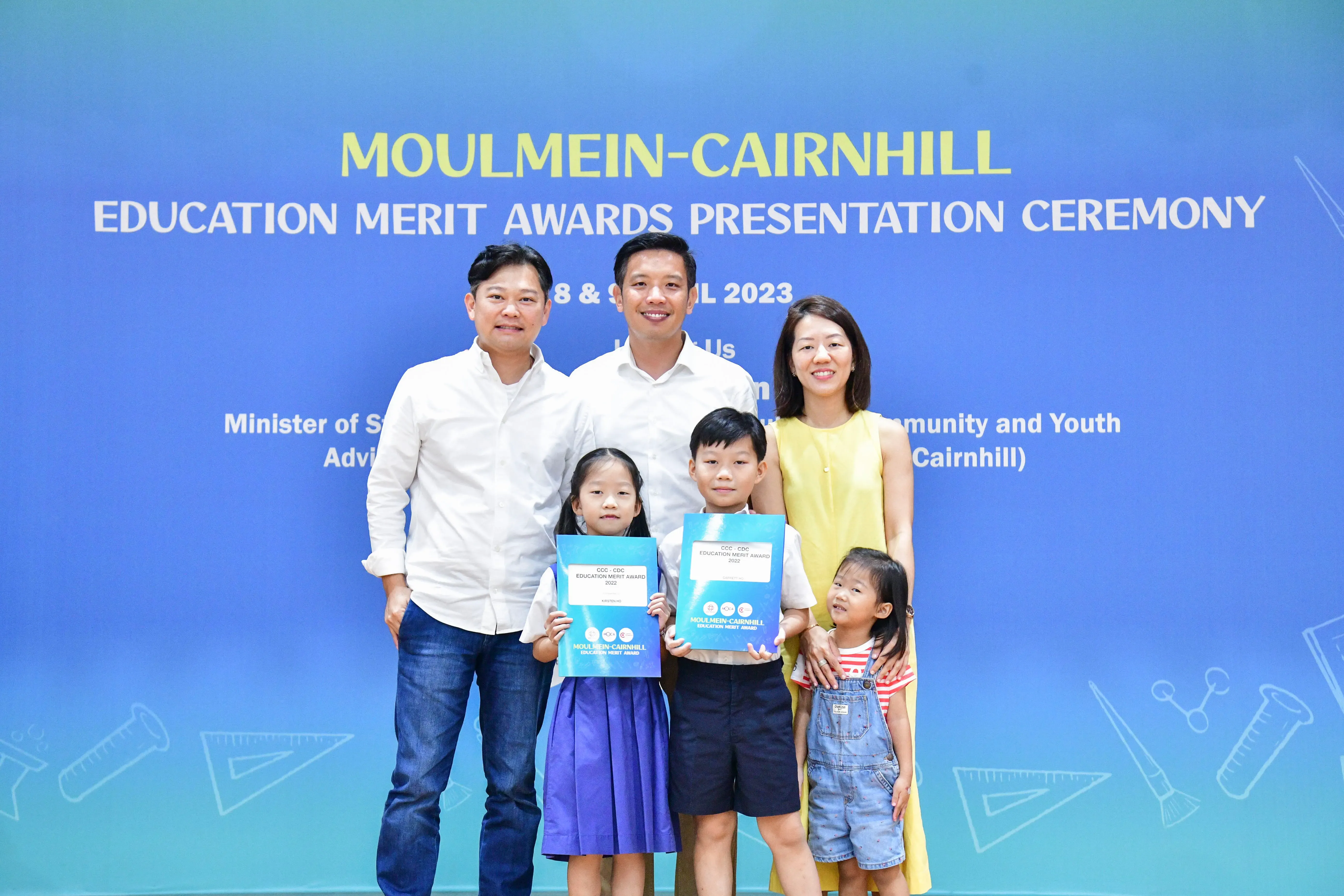 Two adults and three children pose at the Moulmein-Cairnhill Education Merit Awards Ceremony, two kids holding award certificates.