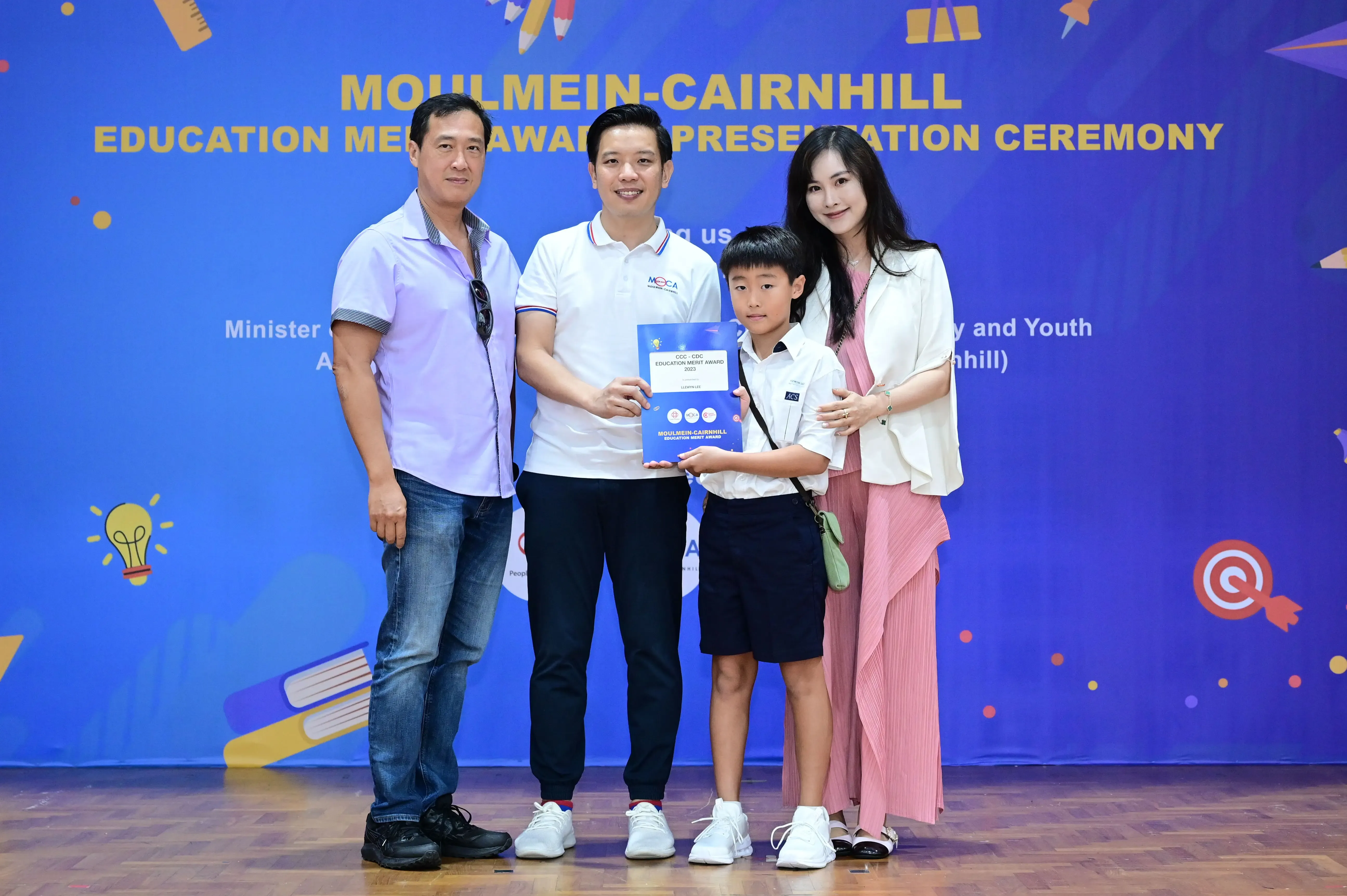 Three adults and a young boy pose holding a certificate at the Moulmein-Cairnhill Education Merit Award ceremony.