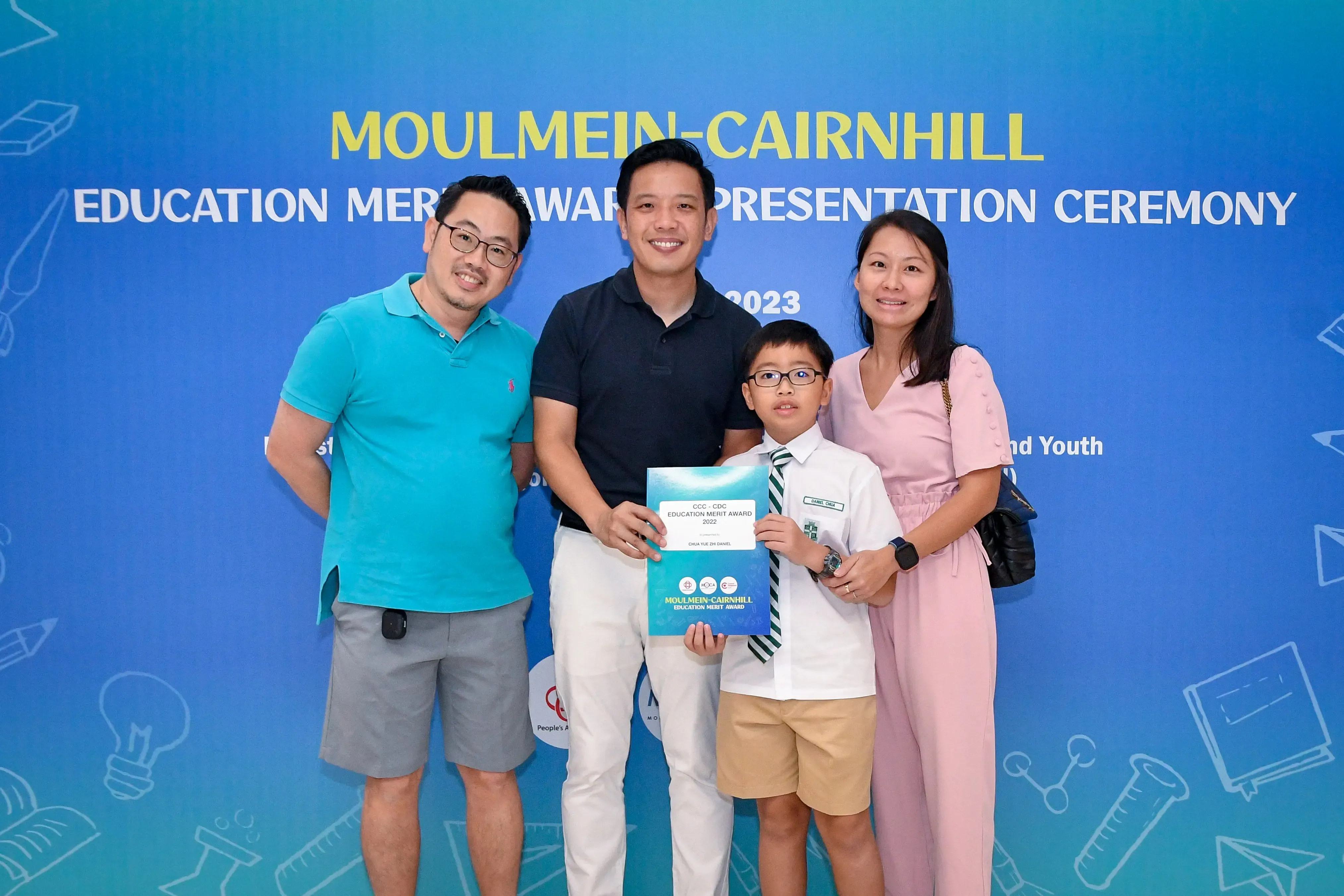Three adults and a boy holding an award certificate pose before a Moulmein-Cairnhill Education Merit Award 2023 backdrop.