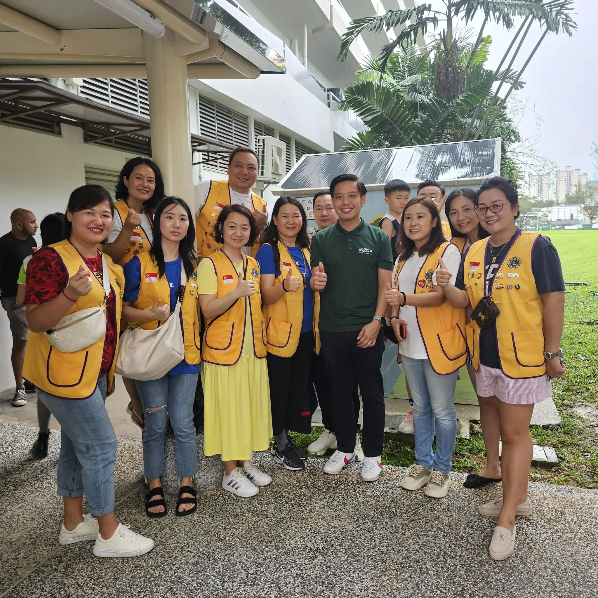 A group of people wearing yellow Lions Club vests pose outdoors giving thumbs up near a building with greenery.