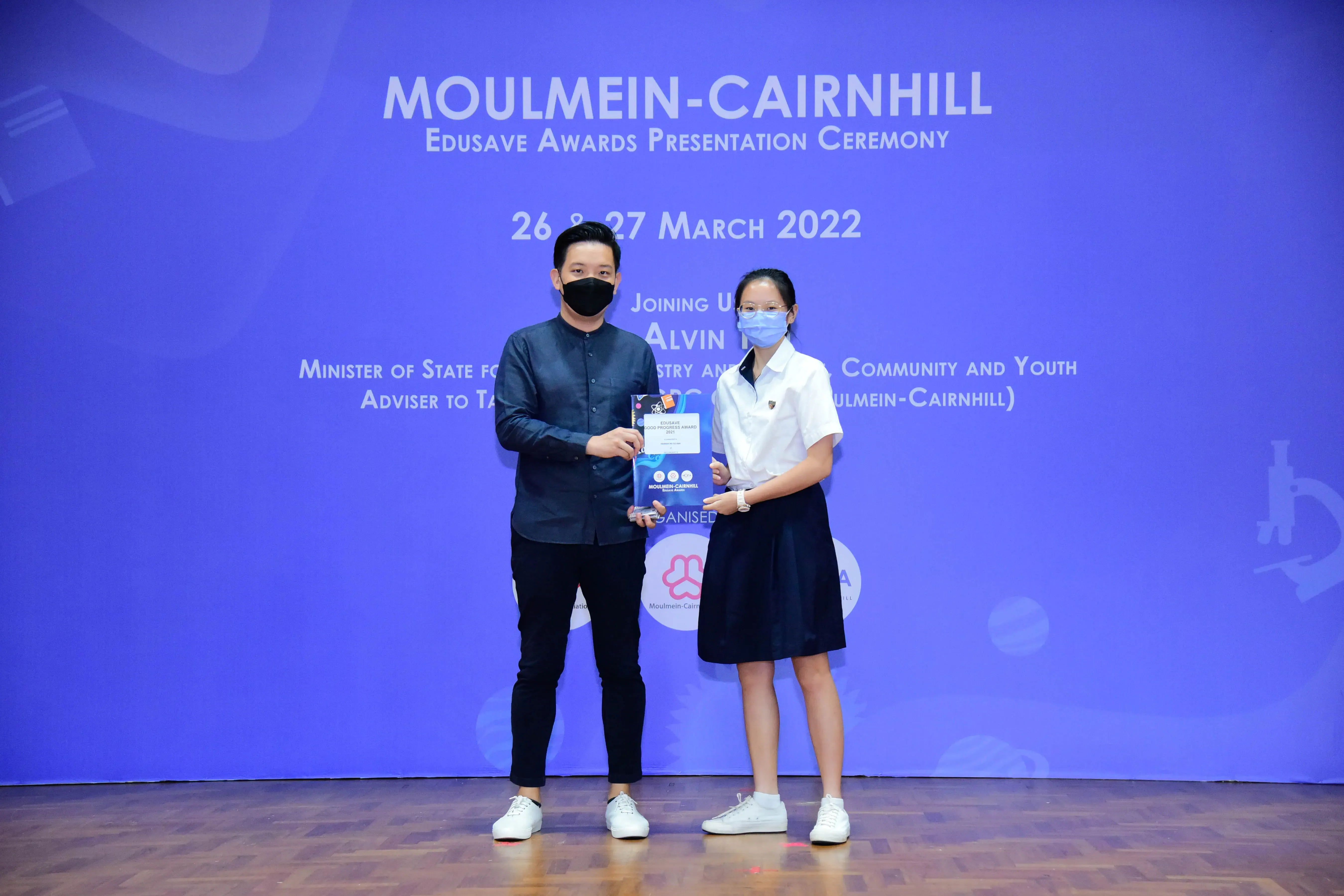 Two masked people hold an award on stage at the Moulmein-Cairnhill Edusave Awards Presentation Ceremony, March 2022.