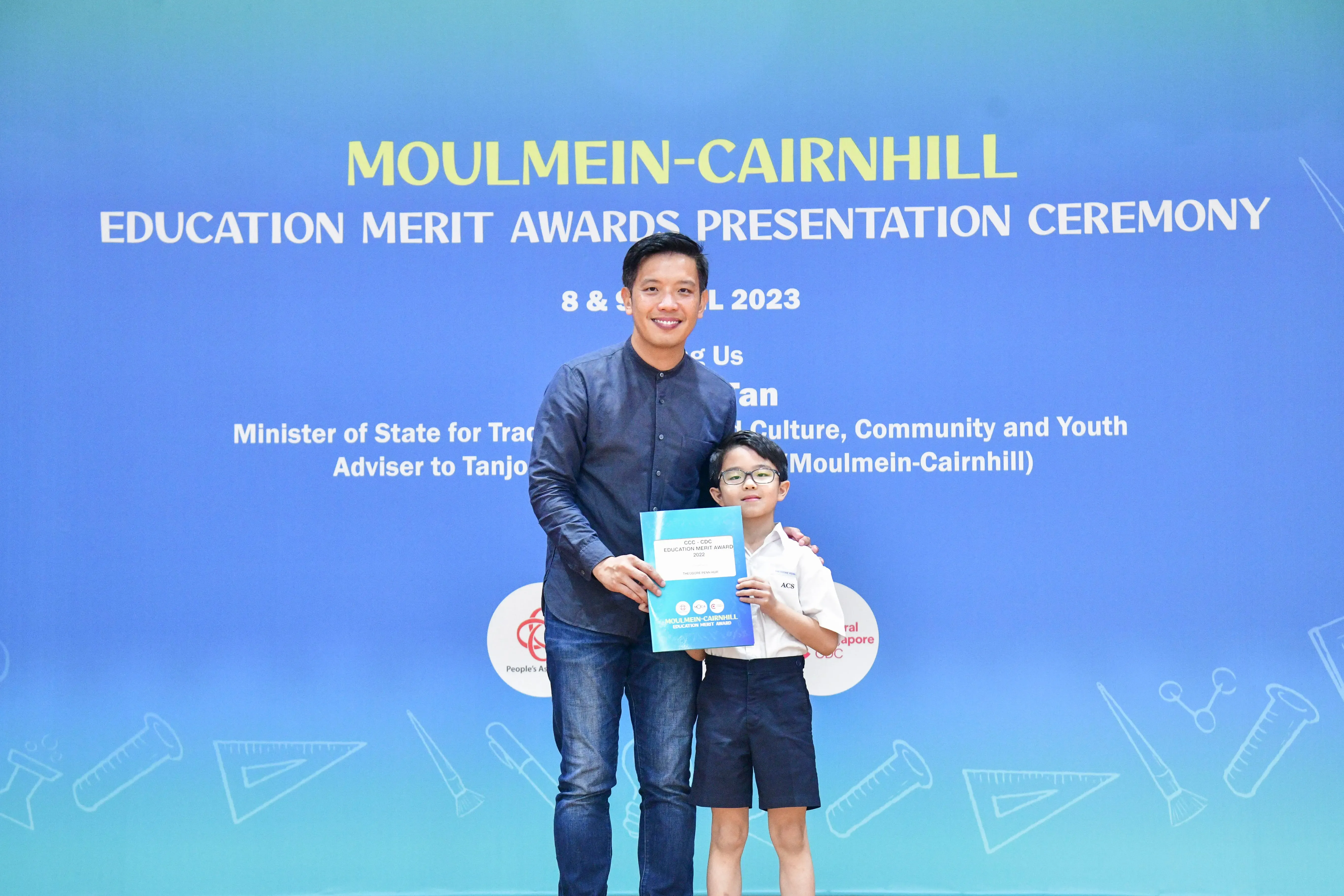 A man and a young boy holding a certificate pose before a Moulmein-Cairnhill Education Merit Awards 2023 backdrop.