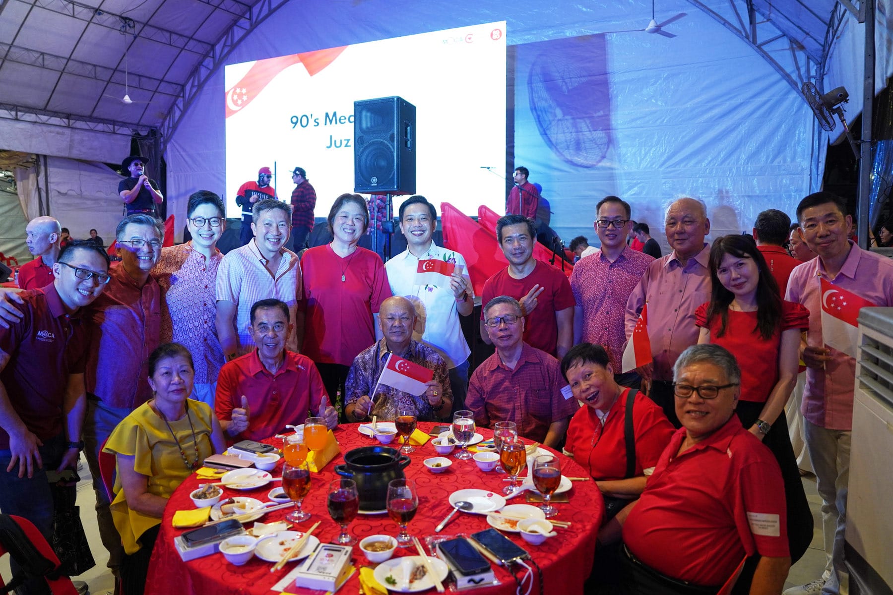 Group of people in red attire posing at a Singapore National Day celebration dinner, holding small Singapore flags.